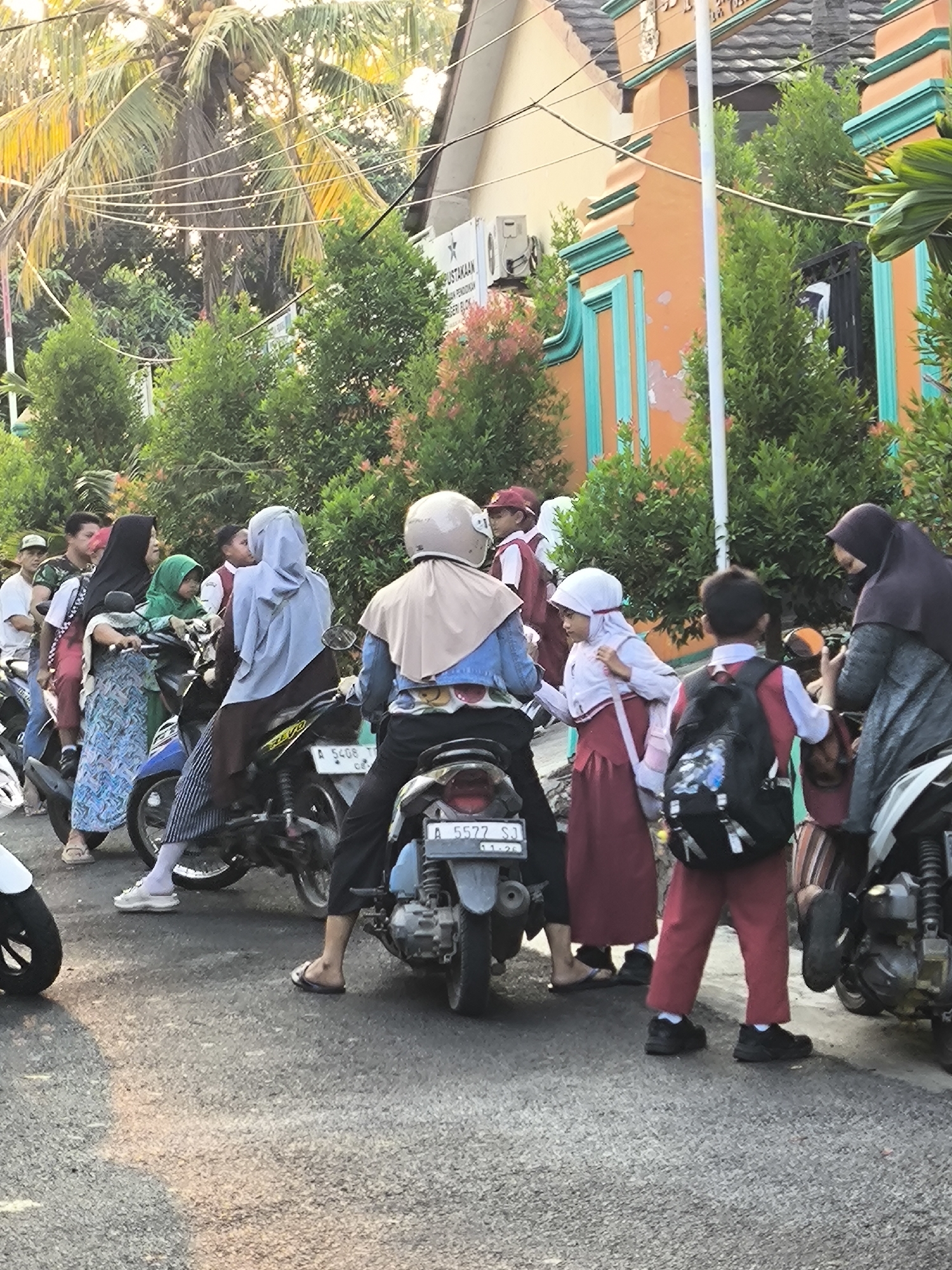 In Cilegon, Indonesia, on a paved street lined with green bushes and trees, a group of people, many wearing school uniforms, are gathered around motorcycles. The background features a building with orange and teal accents, suggesting a public or educational institution. Several individuals are seated on motorcycles, while others stand nearby. Two children in school uniforms, one with a pink backpack, are interacting. The scene appears to be during daylight hours, with soft sunlight illuminating the area. Some individuals are wearing headscarves, indicating a local cultural context. The overall atmosphere suggests a common daily commute or school pick-up.