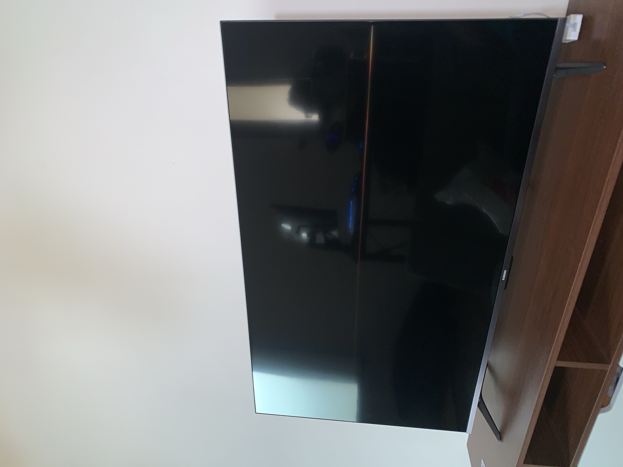 A wide, flat-screen television set with a black frame and stand is placed on a brown wooden media console. The television screen is dark and reflective, showing a distorted reflection of the room, including what appears to be a person with a camera and some blue lights. The media console has two open shelf compartments. The background is a plain white wall. The scene appears to be indoors, possibly in a living room. There is no indication of the location being in Kualadua, Indonesia.