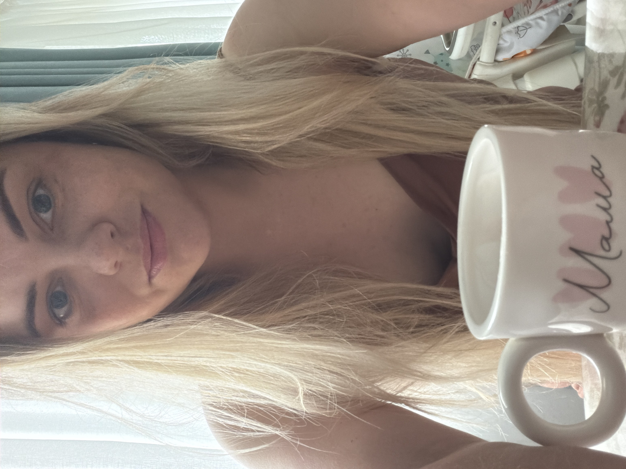 A woman with blonde hair is holding a white mug with the word "Mama" written in script and accented with pink flowers. She is looking at the camera and smiling slightly. Behind her, there is a window with sheer white curtains and a baby chair. The location of the image is unclear and there is no information to associate it with the city of Votkinsk, Russia.