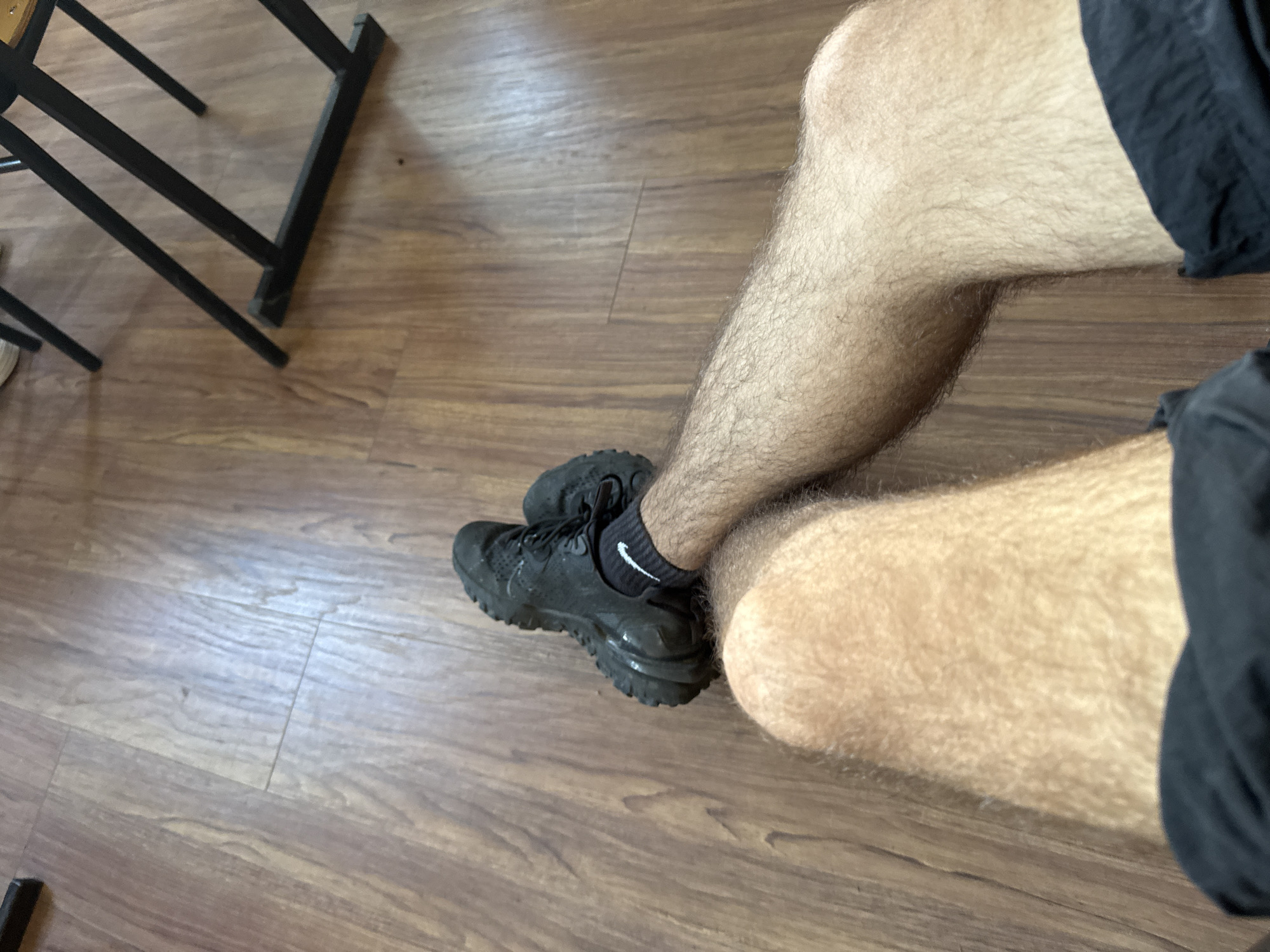 The image shows a close-up view of a person's legs and feet, resting on a wooden floor. The person is wearing black athletic shoes with white socks that have a Nike swoosh logo. The floor is made of light brown laminate wood flooring, with visible grain patterns and seams between planks. In the background, a portion of a black metal structure, possibly a chair or table leg, is visible, indicating an indoor environment. The lighting suggests it is daytime, with some natural light reflecting off the floor. The scene is devoid of any specific indicators placing it in Sofia, Bulgaria, or any other identifiable urban location beyond it being indoors.