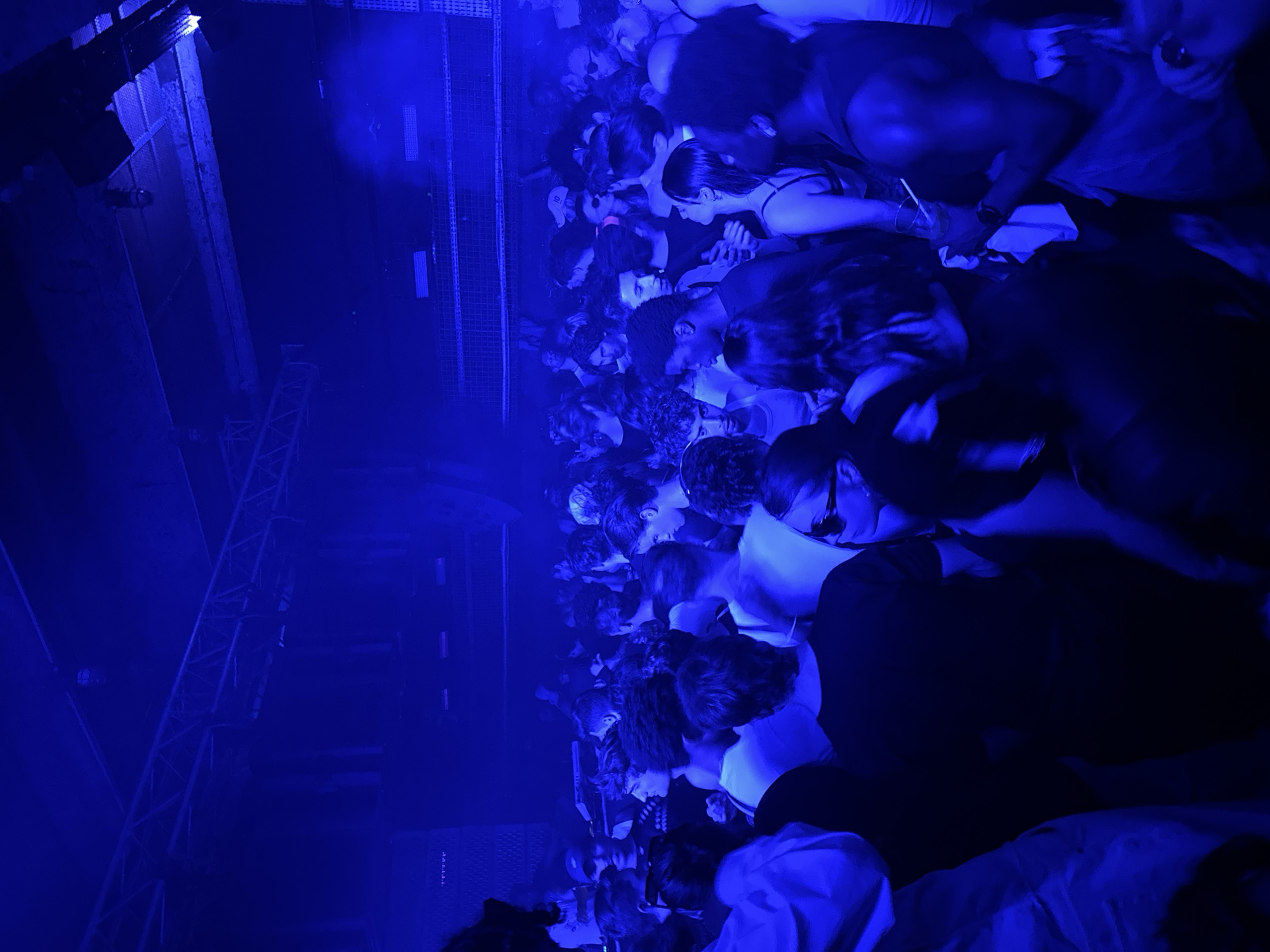 The image captures a crowded indoor event, likely a club night or concert, viewed from an elevated perspective looking down into the audience. The entire scene is drenched in a deep, pervasive blue light, creating a moody and energetic atmosphere.
A dense mass of people fills the frame, with numerous heads and shoulders visible. Attendees are standing closely together, many appearing to face forward, suggesting a central stage or performance area is out of view. Their clothing appears casual, typical for a night-time social gathering. Some individuals are seen holding drinks with straws.
On the left side of the image, substantial metallic truss structures are prominent, indicative of event staging or industrial architecture. These structures appear to support lighting equipment or speakers. A gridded wall or paneling runs along the edge, separating the crowd area.
The activity taking place is a large-scale social gathering, such as a music event or party, occurring at night. The artificial blue lighting is the primary light source, defining the visual tone of the scene. No discernible text is visible within the image. This event is set in Paris, France.