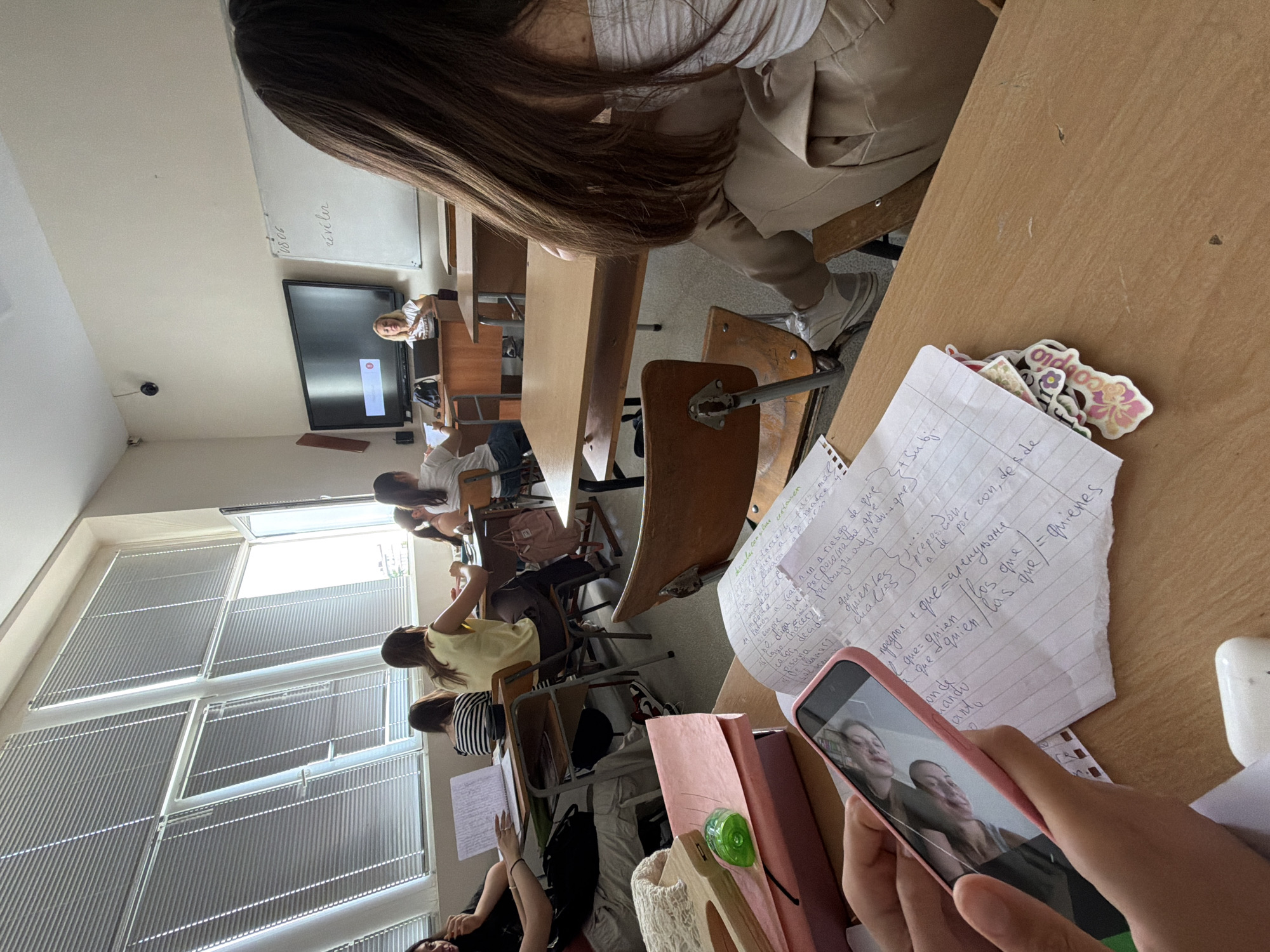 This image captures an indoor scene, likely a classroom in Burgas, Bulgaria, given the context. Several young women are seated at desks, facing forward. A female instructor stands at the front of the room, near a large television screen and a whiteboard. The room features large windows with blinds, and the desks are made of wood. In the foreground, a person's hands hold a pink smartphone, displaying a reflection of two faces. Nearby, there are notebooks with handwritten notes, a green pencil sharpener, and some colorful stickers. The overall atmosphere suggests a learning environment during a lesson.