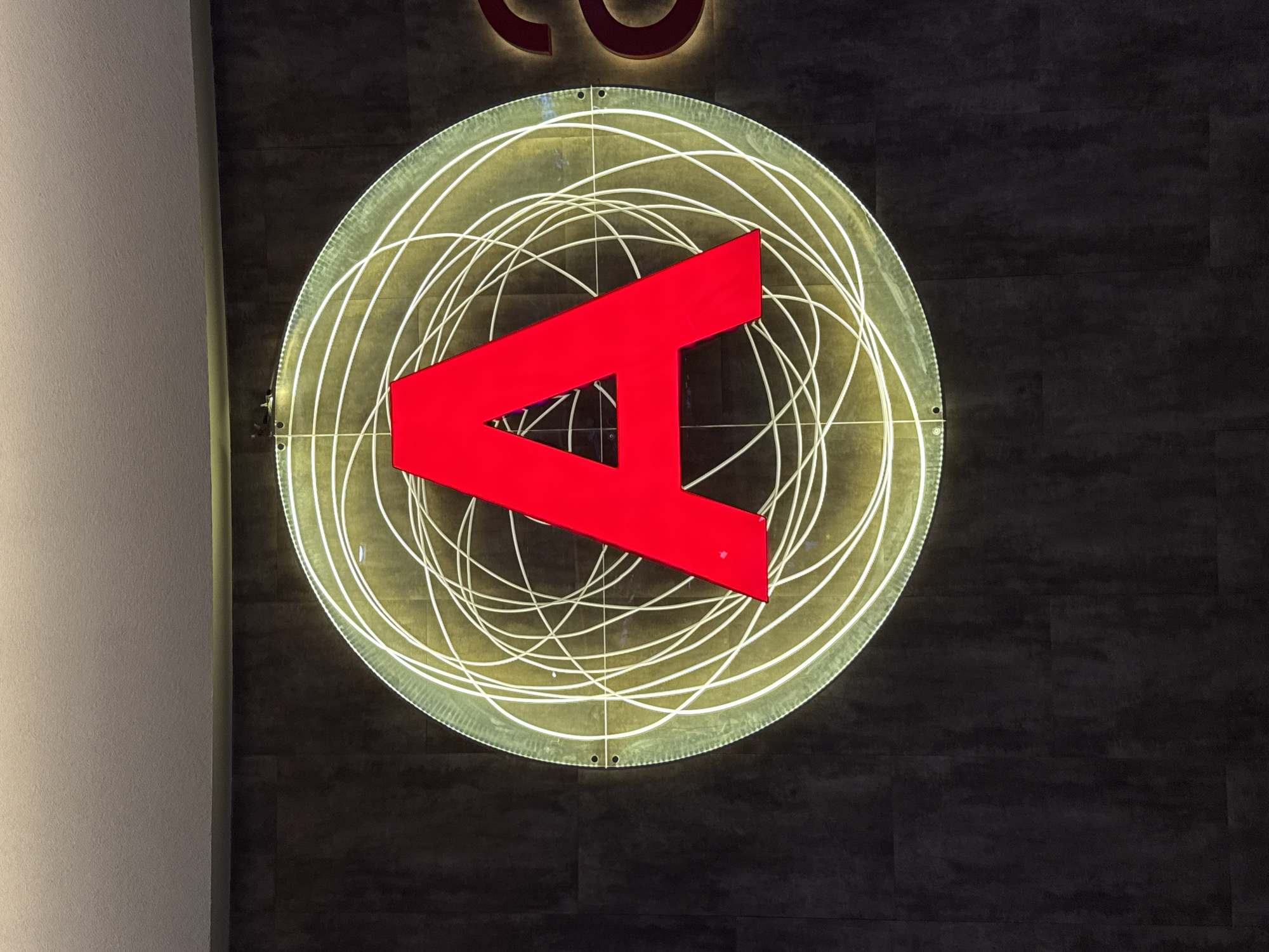 The image shows a circular logo mounted on a dark, textured wall, likely inside a building. The logo consists of a red stylized "A" shape inside a series of concentric, glowing white circles. The circles are not perfectly aligned, giving a somewhat chaotic or dynamic feel. The logo appears to be illuminated from behind, creating a soft, glowing effect. The upper part of another logo (maybe lettering) can be seen above. A white wall is visible on the left edge of the image, suggesting the logo is mounted near a corner. The scene appears to be indoors, in a modern or corporate environment. Location not verified.