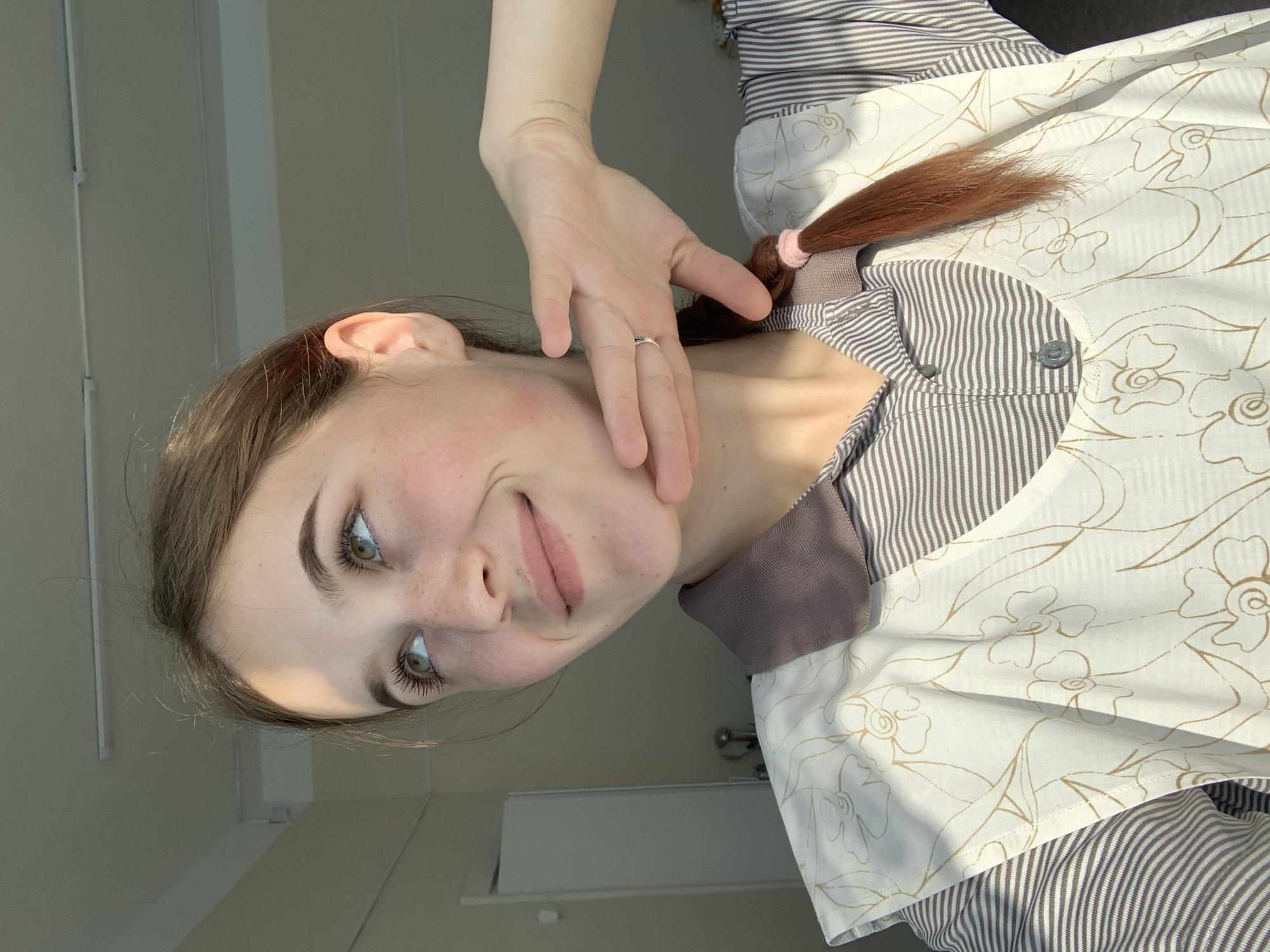 A young woman is shown in a medium close-up, slightly angled, indoors. The location cannot be determined as Фоки, Russia from the available image. She is fair-skinned with freckles and light brown hair pulled back into a ponytail with a pink hair tie. Her hand is raised to her face, adorned with a ring on one finger. She wears a striped shirt with a floral-patterned apron over it. The background is a neutral wall, with some light shining in from the left.