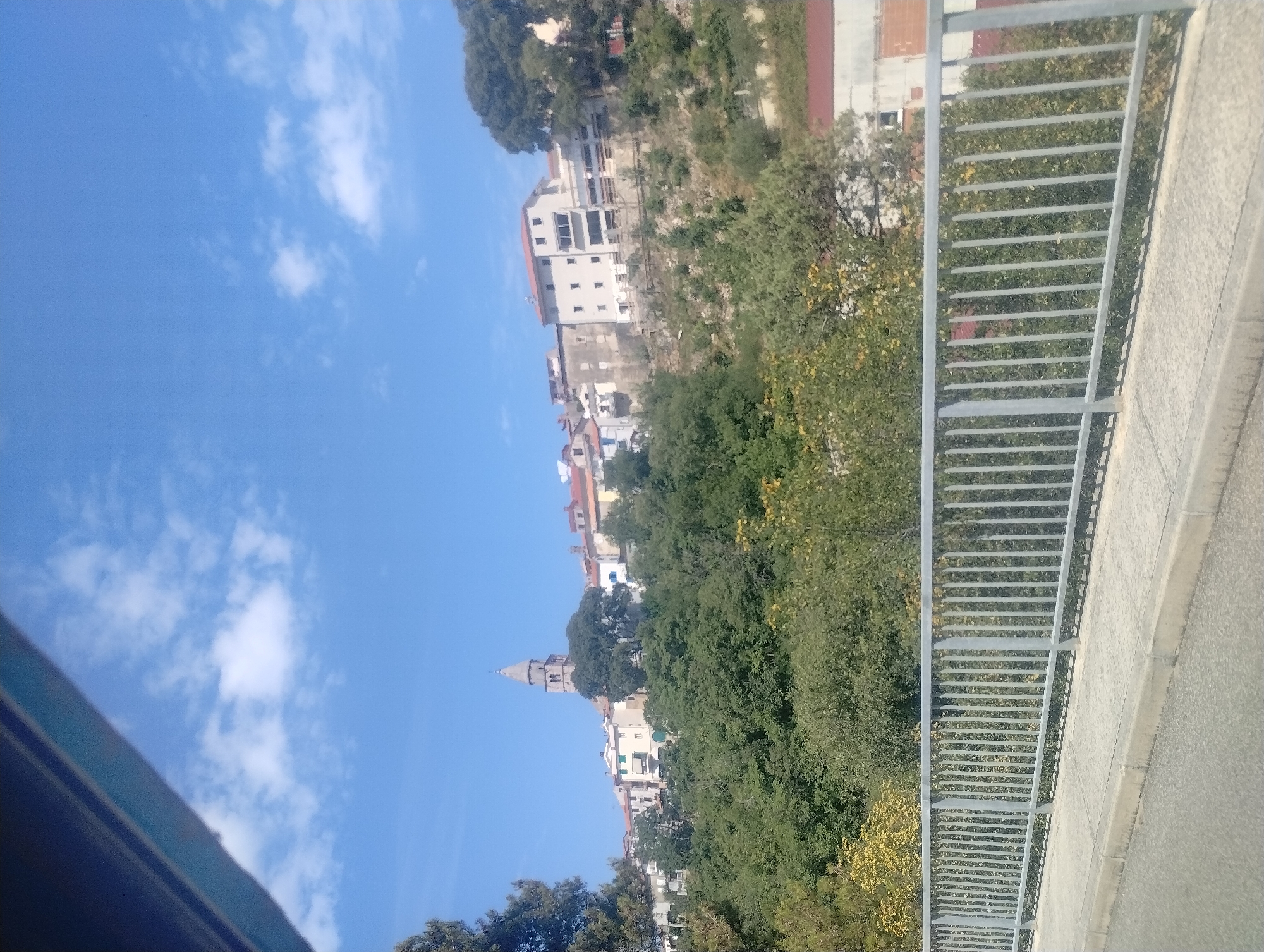 A medium-shot image captures a hillside town under a clear blue sky with scattered white clouds. In the foreground, a metal railing with vertical bars runs horizontally across the frame, defining a walkway or bridge. Beyond the railing, lush green trees cover the lower slope of the hill.
The hillside itself is dotted with buildings, varying in color and architectural style, with red-tiled roofs being a common feature. A prominent, tall, bell tower with a pointed top stands out amongst the buildings, suggesting a church or cathedral. The town appears to be situated on a slope overlooking a body of water, though the water itself is not visible in this frame. The overall scene depicts a picturesque Mediterranean town. The location is identified as Općina Vrbnik, Croatia.