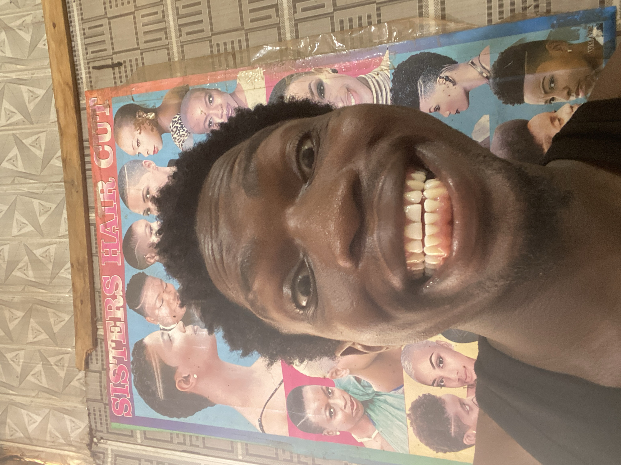 The image is an extreme close-up of a person's face superimposed over a colorful poster. The poster, which appears to be a flyer for a hair salon called "SISTER'S HAIR CUT," features numerous images of people, predominantly women, showcasing various hairstyles. The background of the poster is a vibrant blue, with some sections of pink and other colors. The foreground of the image is dominated by the face of the person, whose head is tilted and positioned in front of the poster. Their eyes are looking directly at the camera, and they have a wide, toothy grin. Their dark, curly hair is visible in the frame. The overall impression is one of a candid, slightly humorous portrait. The setting appears to be indoors, possibly a home or a shop, with a patterned tiled wall visible behind the poster. There are no clear indicators of the time of day or weather. The visible text on the poster reads "SISTER'S HAIR CUT". The location context of Kuson, Nigeria is provided.