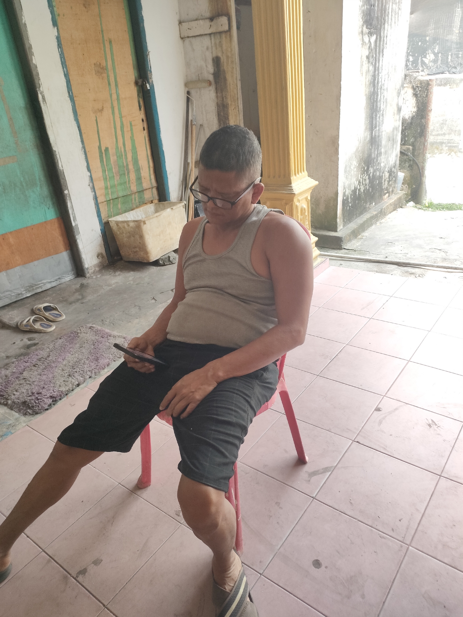 A man is sitting on a red plastic chair, facing left, in what appears to be an outdoor or semi-outdoor area with tiled flooring. He is wearing a grey tank top and black shorts, with glasses and a short haircut. He is looking down at his mobile phone, which he holds in his right hand. To his left, on the tiled floor, is a small, dark-colored mat with a pair of light-colored flip-flops on top of it. In the background, there are wooden doors with green and brown paint, a white plastic container, a fluted yellow column, and a weathered, light-colored wall. The scene suggests a casual, everyday setting, possibly in a residential or commercial area of Pekanbaru, Indonesia, given the architectural elements and common materials.