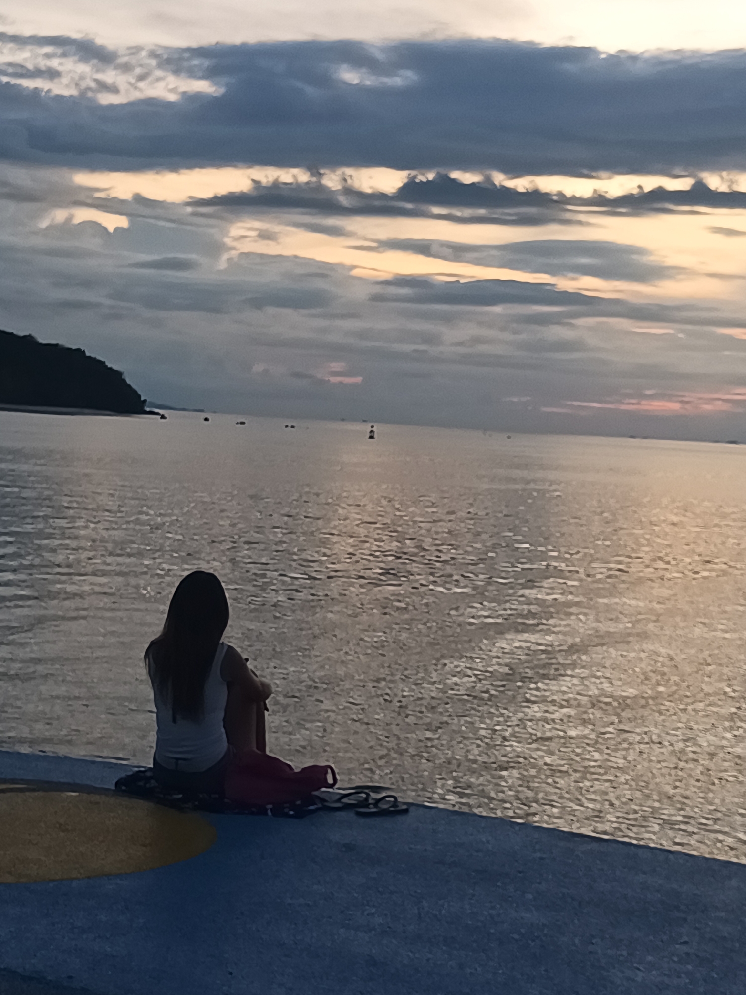 A solitary woman, seen from behind, sits on a paved surface overlooking a vast expanse of water. She is dressed in a white tank top and shorts, with her legs drawn up towards her chest. Beside her rests a pink bag and some dark chain-like objects. The water reflects the twilight sky, creating a shimmering effect. In the distance, a silhouetted landmass, possibly an island, juts out from the left side of the frame, with a few small boats or buoys dotting the horizon. The sky is filled with dramatic clouds, tinged with the soft hues of sunset, suggesting either dawn or dusk. The overall mood is one of quiet contemplation or solitude. The location is identified as Koh Phangan, Thailand.