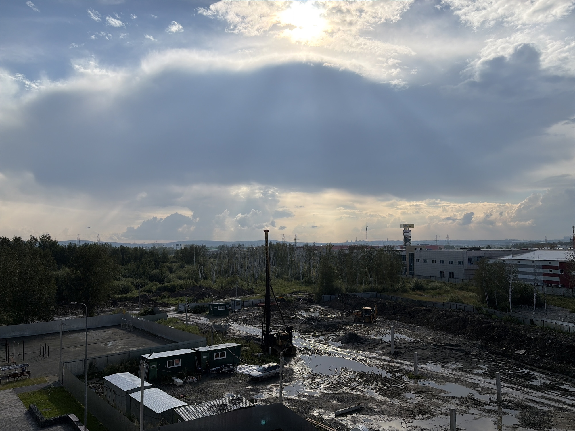 The image captures an overcast sky with the sun peeking through dramatic clouds, casting a diffuse light over a construction site in Yekaterinburg, Russia. The scene depicts a muddy, waterlogged area with tire tracks and piles of dirt, indicating ongoing earthwork. A large drilling rig stands prominently in the center of the site, accompanied by an orange excavator in the background. Several temporary modular buildings are present, including green structures with windows. A paved area with a fence and some exercise equipment is visible in the foreground, adjacent to a building with a silver corrugated metal roof. In the distance, industrial buildings and power lines are discernible, suggesting the location is on the outskirts of the city. The overall atmosphere is one of industrial activity under a dynamic sky.