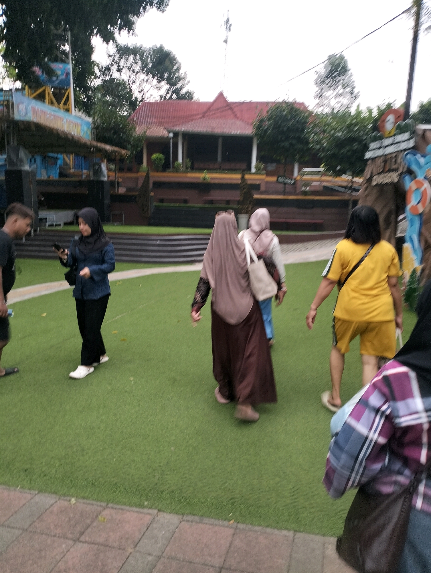 The image depicts an outdoor scene, possibly at a recreational area. In the foreground, a person wearing a purple and white plaid shirt and a dark bag is partially visible on the right. Moving towards the center, several individuals are walking on a green, artificial turf surface. One woman, seen from behind, wears a yellow t-shirt and yellow shorts, with a dark strap across her chest. To her left, a person in a long brown garment and a tan headscarf is walking away, carrying a light-colored bag. Another person in a light pink headscarf is walking ahead of them. On the left side of the image, a woman in a blue long-sleeved shirt and black pants, wearing a black headscarf, is looking at her phone. A person in a black t-shirt is visible to her left. In the background, there is a building with a red roof, trees, and what appears to be playground equipment or an attraction with blue and yellow structures. The environment seems to be a public space, possibly a theme park or a park with recreational facilities, located in Karangbanjar, Indonesia, as suggested by the context.