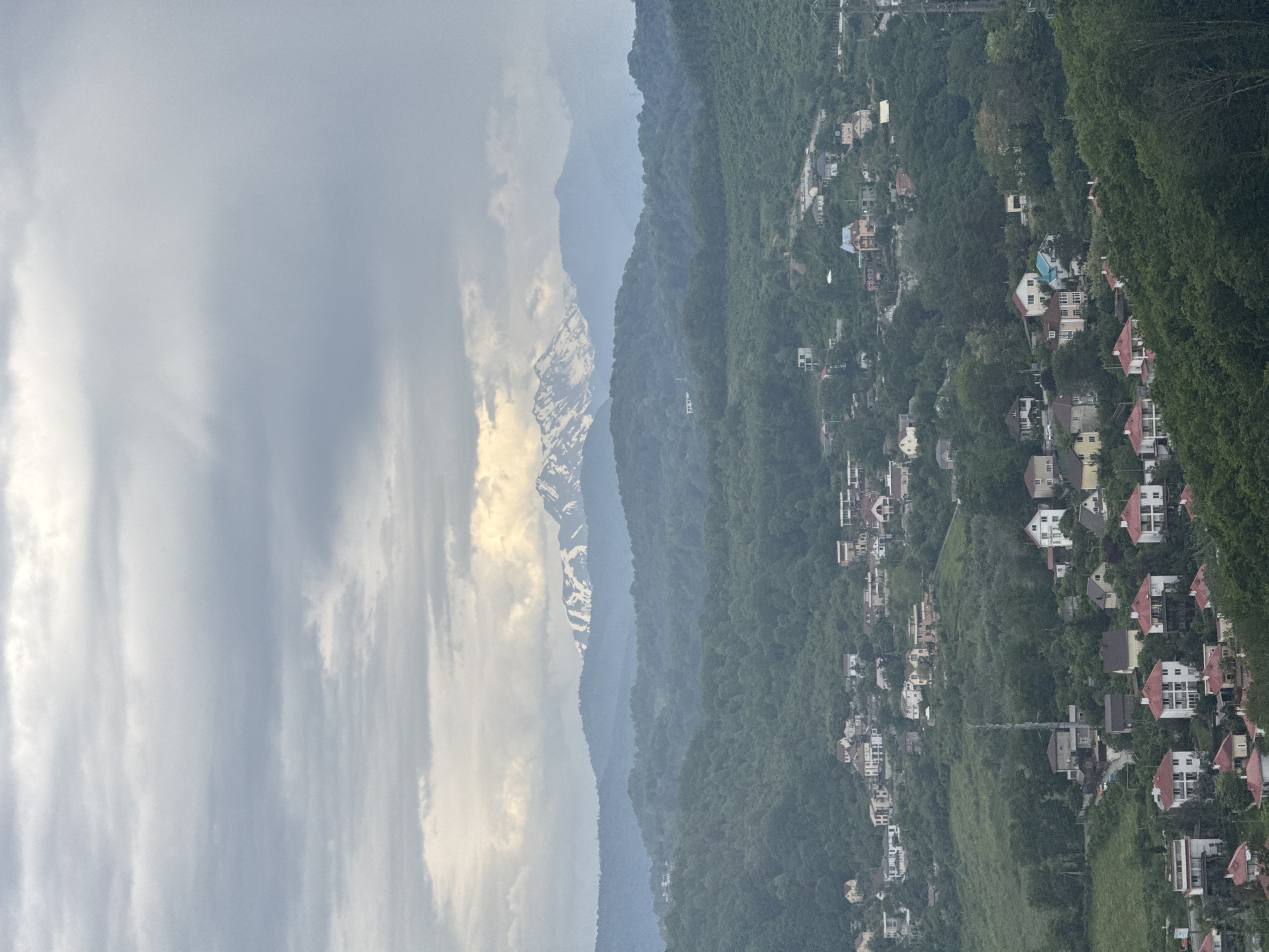 The image shows a mountain range, a city nestled on a mountain side, and a cloudy sky. The mountain range is in the background, with some of the peaks covered in snow. In the foreground is a city on a steep mountainside, partially obscured by foliage. Houses are densely packed together, with a variety of roof colors. The sky is heavily clouded.
The location is likely the city of Sochi, Russia, as it is known for its mountainous terrain, the Black Sea coast, and surrounding vegetation.