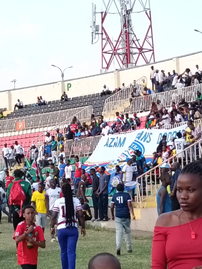 The image displays an outdoor stadium scene in Nairobi, Kenya, depicting a public gathering consistent with a sports event. The perspective is from ground level, looking towards the stadium's tiered seating and the sky.
In the upper background, a large red and white cellular communications tower is prominently visible against a light sky. Below it, the stadium structure features a concrete wall with a green "P" symbol, followed by multiple tiers of seating. These seats are predominantly dark gray, interspersed with sections of red and green seating. White railings delineate the different seating areas. Numerous individuals, primarily adults, occupy various sections of the stands, some seated, others standing.
At the mid-ground, a large white banner is stretched across a lower section of the stands. Blue text on the banner is partially visible, clearly showing "AZAM" and "ANS DODO," with "TBA" also discernible. Flags, including some green and white, are visible among the crowd in the stands on the left.
The foreground and mid-ground are populated by multiple individuals walking on a grassy or dirt surface, consistent with a perimeter path or the edge of a playing field. Notable persons include:
* A young male in a red short-sleeved shirt, holding a brown bottle in his left hand, looking forward.
* A woman with long, dark braided hair, wearing a white, red, and black striped jersey with the number "11" on the back, and blue pants, walking away from the camera.
* A man in a dark blue short-sleeved shirt with the number "41" on the back and blue jeans, also walking away.
* Various other individuals, some wearing team-branded apparel, such as a yellow shirt with "QATAR AIRWAYS" and "PSG" logos.
* In the immediate right foreground, a woman wearing an off-the-shoulder red top with a red whistle suspended around her neck is visible, looking towards her right.
* The top of a person's head is partially visible at the bottom center of the frame.
The overall environment indicates an active public event, with both spectators in designated seating and individuals moving at ground level, suggesting either ingress, egress, or activities during an interval.