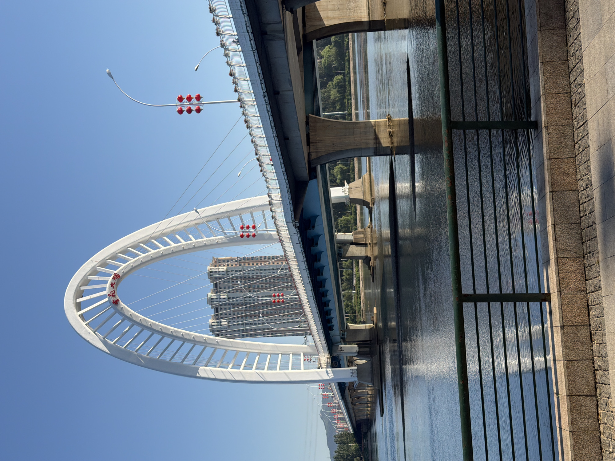 The image shows a wide bridge spanning a body of water, likely a river, in the city of Linghe, China. The bridge features a prominent white archway with red Chinese characters inscribed at its apex. Red lanterns are strung along lampposts on the bridge. A tall, modern building is visible behind the archway. The water reflects the sky and the bridge. In the foreground, a green metal railing separates the viewer from a tiled walkway. The sky is clear and blue, indicating a sunny day.