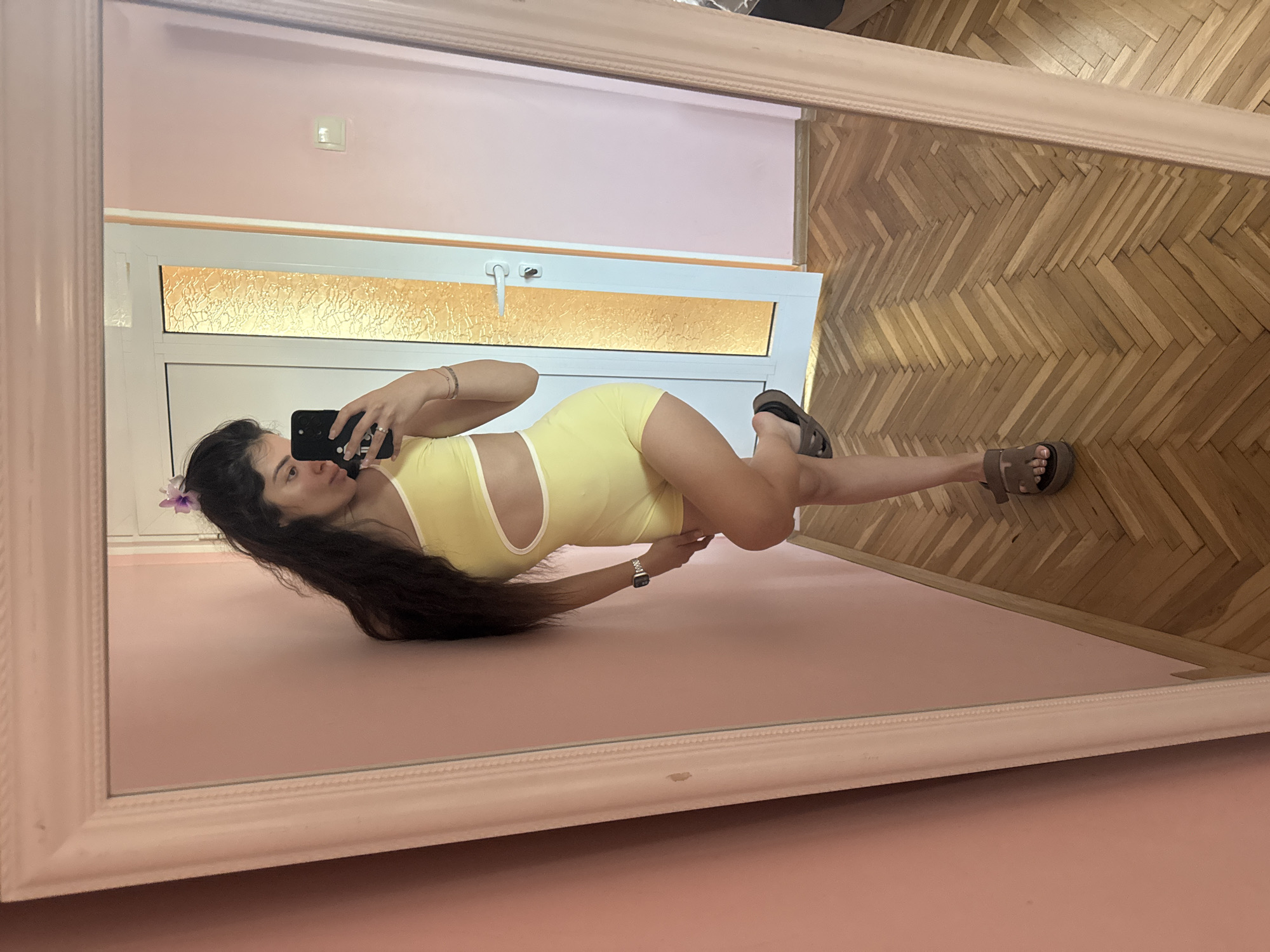 A woman is taking a selfie in a mirror. The mirror shows a reflection of a room with pink walls and a wooden floor. The woman is wearing a yellow outfit and sandals. There is a window in the background. This image might be taken in Plovdiv, Bulgaria, however, there is no irrefutable evidence to support this.
