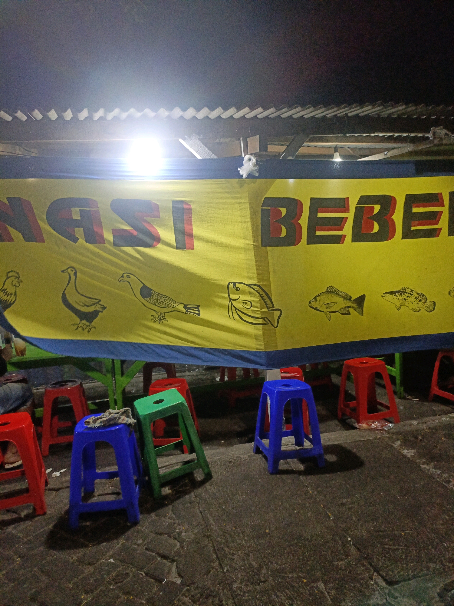 The image depicts an outdoor food stall, likely located in Probolinggo, Indonesia, at night. A bright yellow banner with bold, red-outlined text announces "NASI BEBEK" (duck rice) and features simple illustrations of animals, including a chicken, ducks, and fish. Underneath the banner, various colorful plastic stools are arranged around tables, suggesting a dining area. Some of these stools are red, one is blue, and another is green. The ground is paved with cobblestones. A bright light source, possibly a lamp or floodlight, illuminates the banner from above, creating harsh shadows. The background is dark, indicating nighttime, with a corrugated roof structure visible above the banner. No people are clearly visible or identifiable.