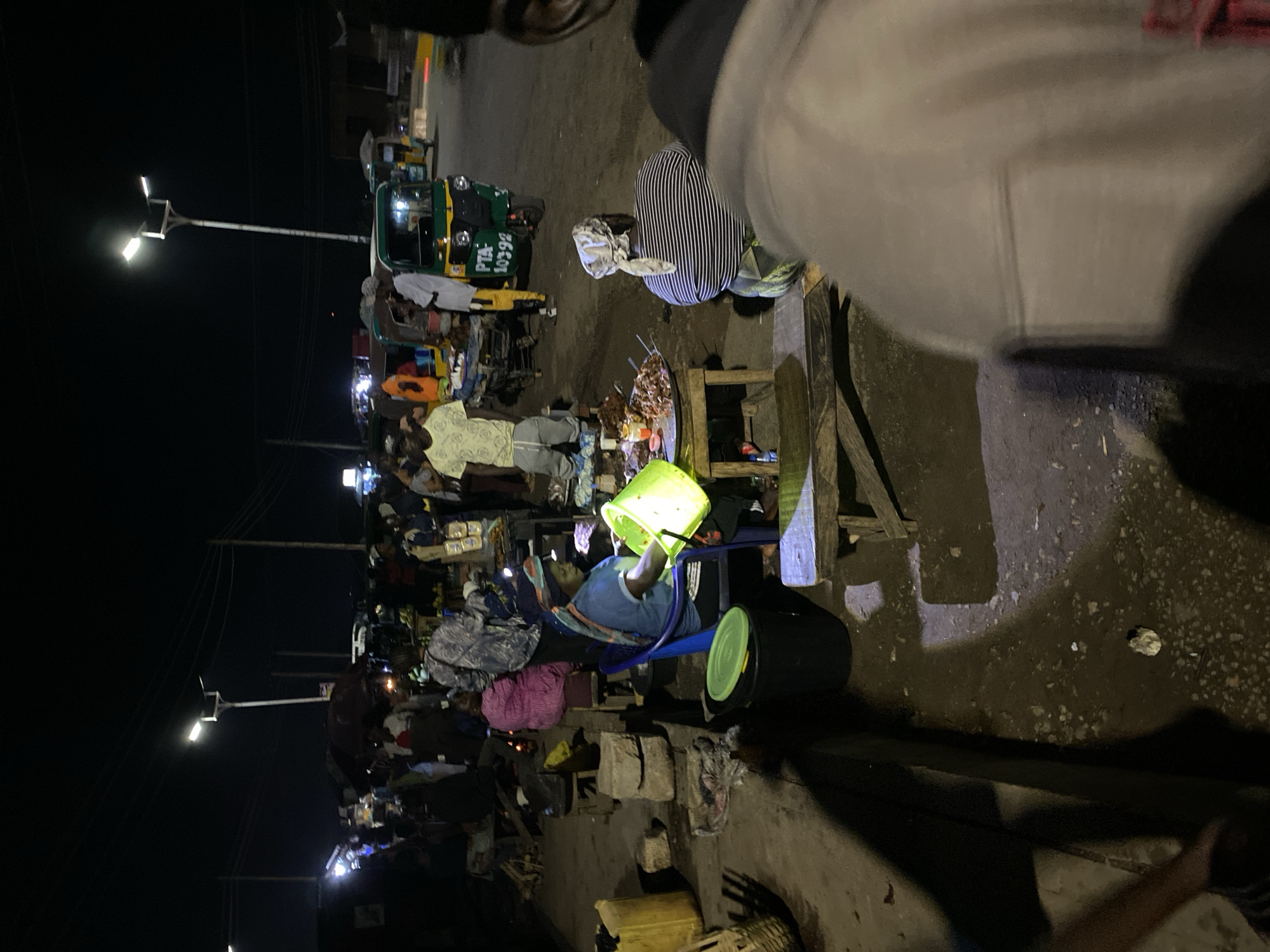 At night in Bukuru, Nigeria, an outdoor market bustles with activity. Several people are gathered around various stalls and vendors. Many people are buying food. A green auto rickshaw (tuk-tuk) is visible near one of the stalls. Streetlights illuminate the scene, casting light onto the market and the surrounding area. There is a mix of individuals, some appearing to be vendors selling goods, while others are potential customers. The ground is paved. A woman sits on a blue chair and appears to be prepping food.