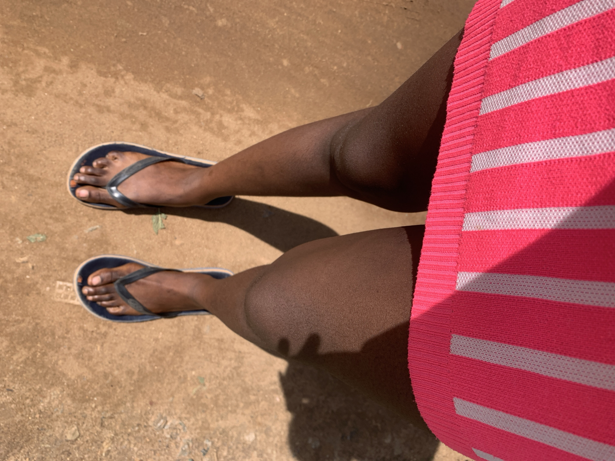 The image presents a downward-facing view of an individual's lower body from the upper thighs to the feet, positioned on an unpaved, reddish-brown earthen surface in Rafin Par, Nigeria. The person has a dark skin tone. The lower edge of a bright pink garment with prominent vertical white stripes, featuring a ribbed hem, is visible at the top of the frame. The individual is wearing dark blue flip-flops with light-colored soles. Strong sunlight illuminates the scene, evidenced by distinct shadows of the legs cast on the ground. The ground surface displays minor textural irregularities consistent with dry soil.