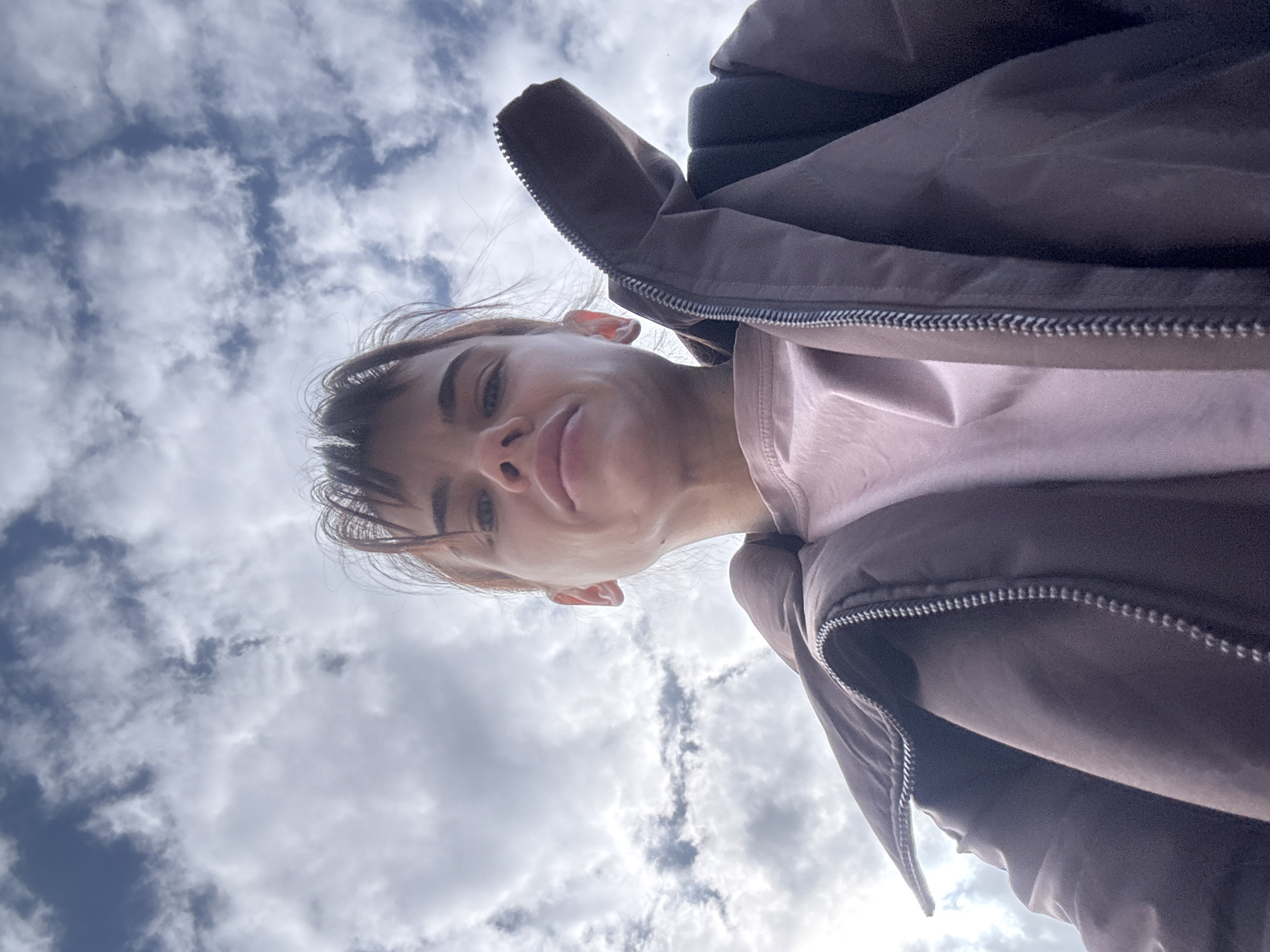 An upward-facing, low-angle shot captures a person against a dynamic, cloudy sky. The primary subject is a young woman, seen from the chin up, with her face dominating the lower-middle portion of the frame. She wears a light pink t-shirt underneath a dark brown or grey puffer jacket, which is partially unzipped. Her hair is dark brown, and a fringe falls across her forehead. She has a serene expression, with a subtle, closed-mouth smile.
The camera's extreme low perspective places the viewer directly beneath the subject, making her appear to look down into the lens. Her face is slightly angled, revealing her left ear and the delicate structure of her jawline. The jacket's hood or collar frames the upper right side of the image, showcasing its fabric texture and visible zipper teeth.
The background is a bright, partially cloudy daytime sky. Patches of deep blue sky are visible through scattered, fluffy white cumulus clouds, suggesting fair weather with some cloud cover. The sunlight diffuses through the clouds, indicating it is daytime. No specific location cues are visible in the image itself, but the provided context places the scene in Rostov-on-Don, Russia. The warm jacket suggests cooler temperatures. There is no visible text in the image.