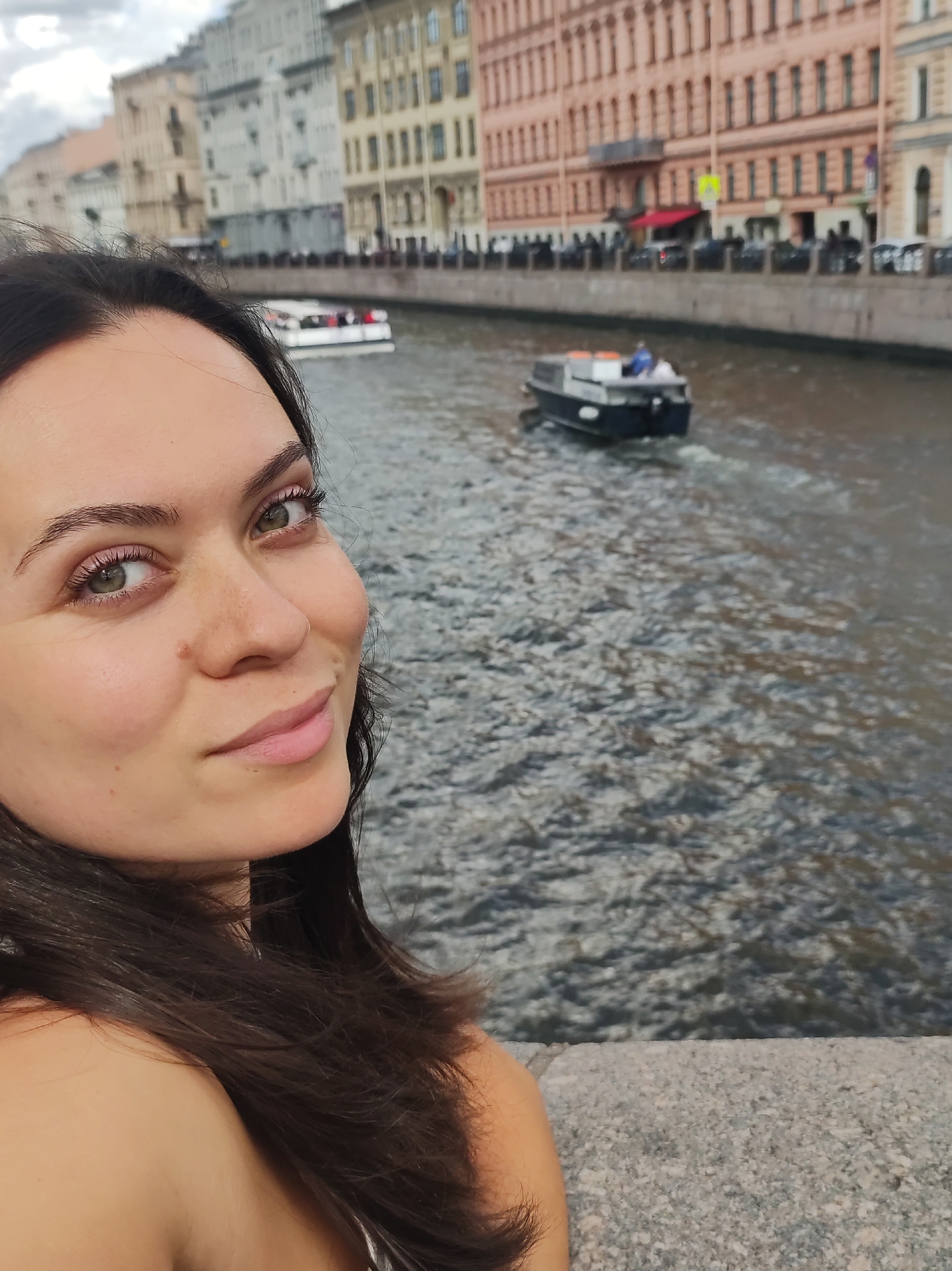 This is a selfie taken by a woman with dark hair and green eyes. She is looking at the camera with a slight smile, with her face positioned in the left portion of the frame. Behind her, a canal runs parallel to a street lined with colorful, multi-story buildings. A boat is visible in the canal, moving away from the camera. The scene appears to be in Saint Petersburg, Russia, given the architectural style of the buildings and the presence of a canal. The sky is overcast, and the water is choppy. The woman is leaning against a stone embankment.