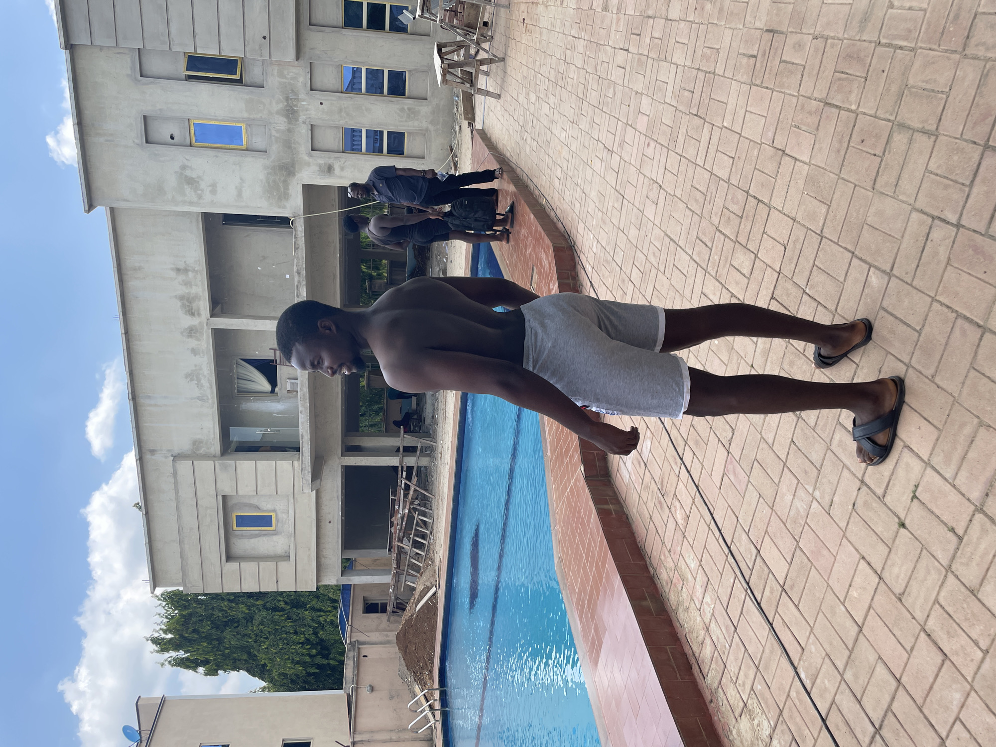 A shirtless man in grey shorts and black sandals stands at the edge of a swimming pool next to a brick-paved walkway. The swimming pool is blue and appears to be outdoors. Behind the man is an unfinished three-story building with scaffolding. A group of people stands behind the man towards the back of the image. The sky is partly cloudy. The location is Akwanga, Nigeria.