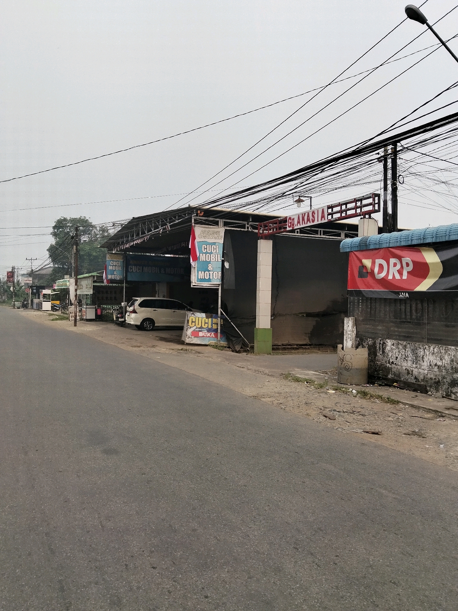 The image shows a roadside view of a car wash and motorbike service business named "ARRIEZ CARWASH" in the city of Arang Limbung, Indonesia. The facade of the business features a dark-colored awning extending over the service area. Several signs are visible, including one with a red and white Indonesian flag and text that reads "CUCI MOBIL & MOTOR" (Car and Motor Wash). Another sign displays the text "DRP". A white MPV is parked within the service area, and a motorbike is parked outside to the left. Overhead, a dense network of electrical wires crisscrosses the sky, supported by utility poles, and a street lamp is visible on the right. The road in front of the business appears to be asphalt and is empty of traffic. The environment is somewhat cluttered with some debris visible along the roadside. The sky is overcast.