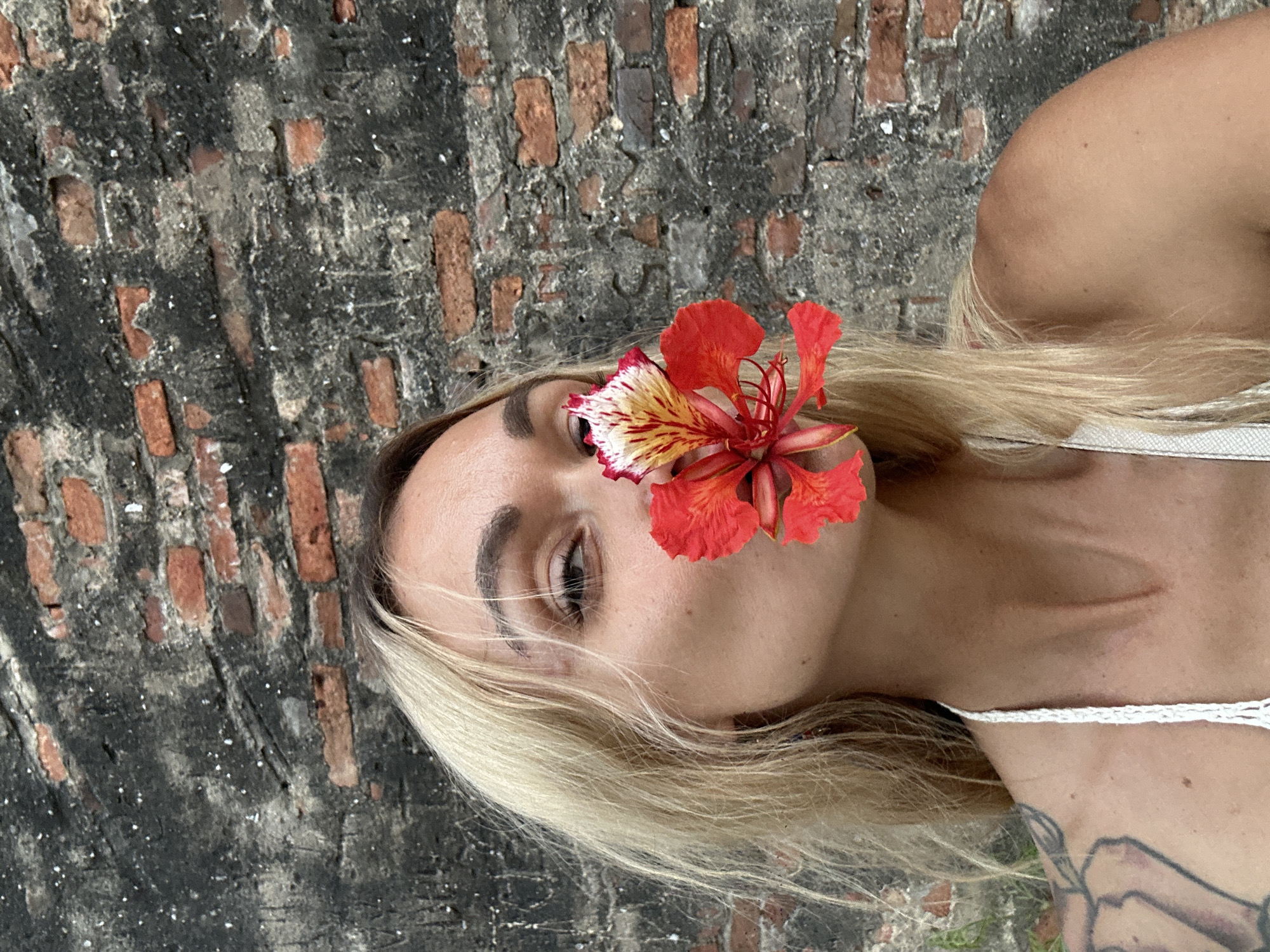 A woman with blonde hair and a red and yellow flower positioned in front of her face is shown in a medium shot against a brick wall. The woman is wearing a white top, and a tattoo is visible on her shoulder. The background consists of an aged brick wall with varying tones of red, grey, and black. The shot appears to be taken outdoors, with some green vegetation at the lower corner. The scene is suggestive of a location that has a mix of natural and constructed elements. No specific evidence to positively ID the location as Phu Thuy Ward, Vietnam can be provided from the image alone.