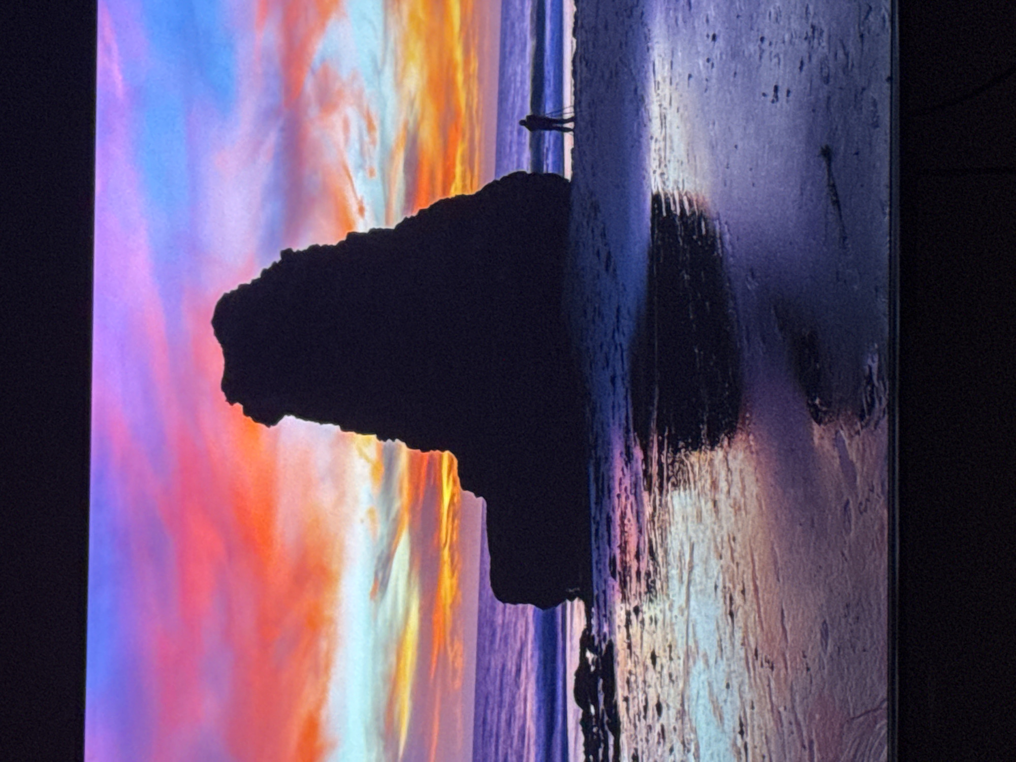 The image depicts a dramatic sunset over a beach scene. A large, dark rock formation dominates the center of the frame, casting a silhouette against a vibrant sky filled with hues of orange, pink, and purple. The ocean stretches out in the background, with gentle waves meeting the shore. In the foreground, the wet sand reflects the colors of the sky, creating a mirror-like effect. A lone figure, possibly a photographer with a tripod, is visible on the beach to the right of the rock formation. The overall atmosphere is serene and picturesque. This scene is not located in Saratov, Russia.