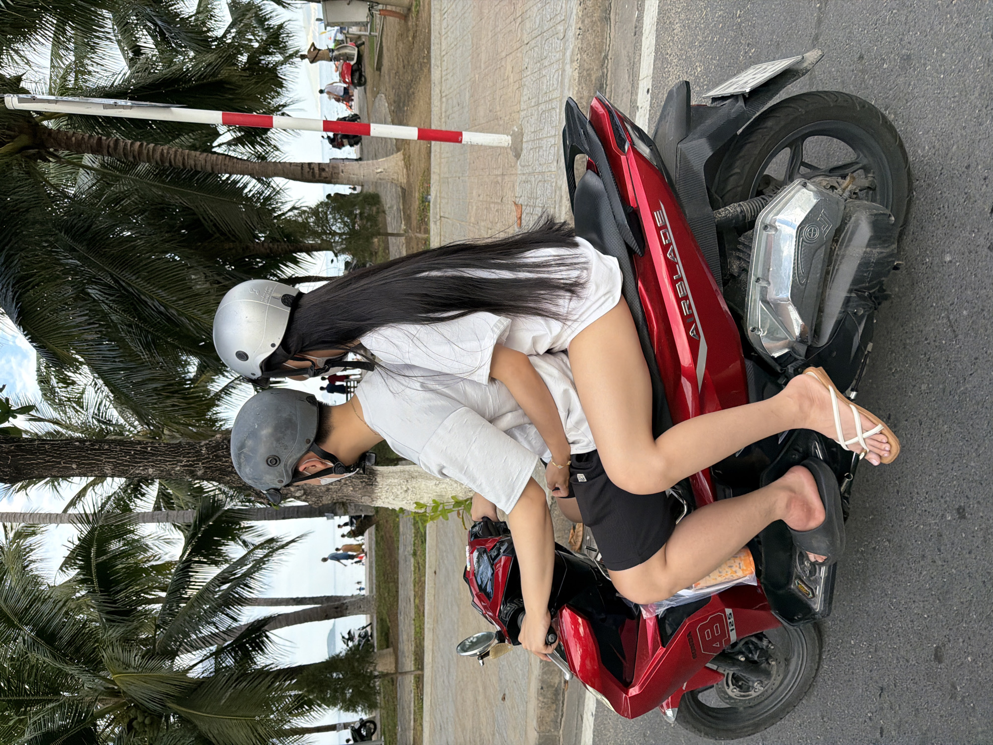 In Nha Trang, Vietnam, a man and a woman ride a red scooter along a paved road lined with palm trees and a pedestrian walkway. The man, wearing a grey helmet and a white t-shirt, drives the scooter, while the woman, with long black hair and a silver helmet, sits behind him, holding onto his waist. She wears a white t-shirt and black shorts, and her legs are visible, one bare and the other covered by a flip-flop. The scooter has "AIRBLADE" printed on its side. In the background, people walk along the beach, and several other scooters are parked. A red and white striped pole stands near the walkway.
