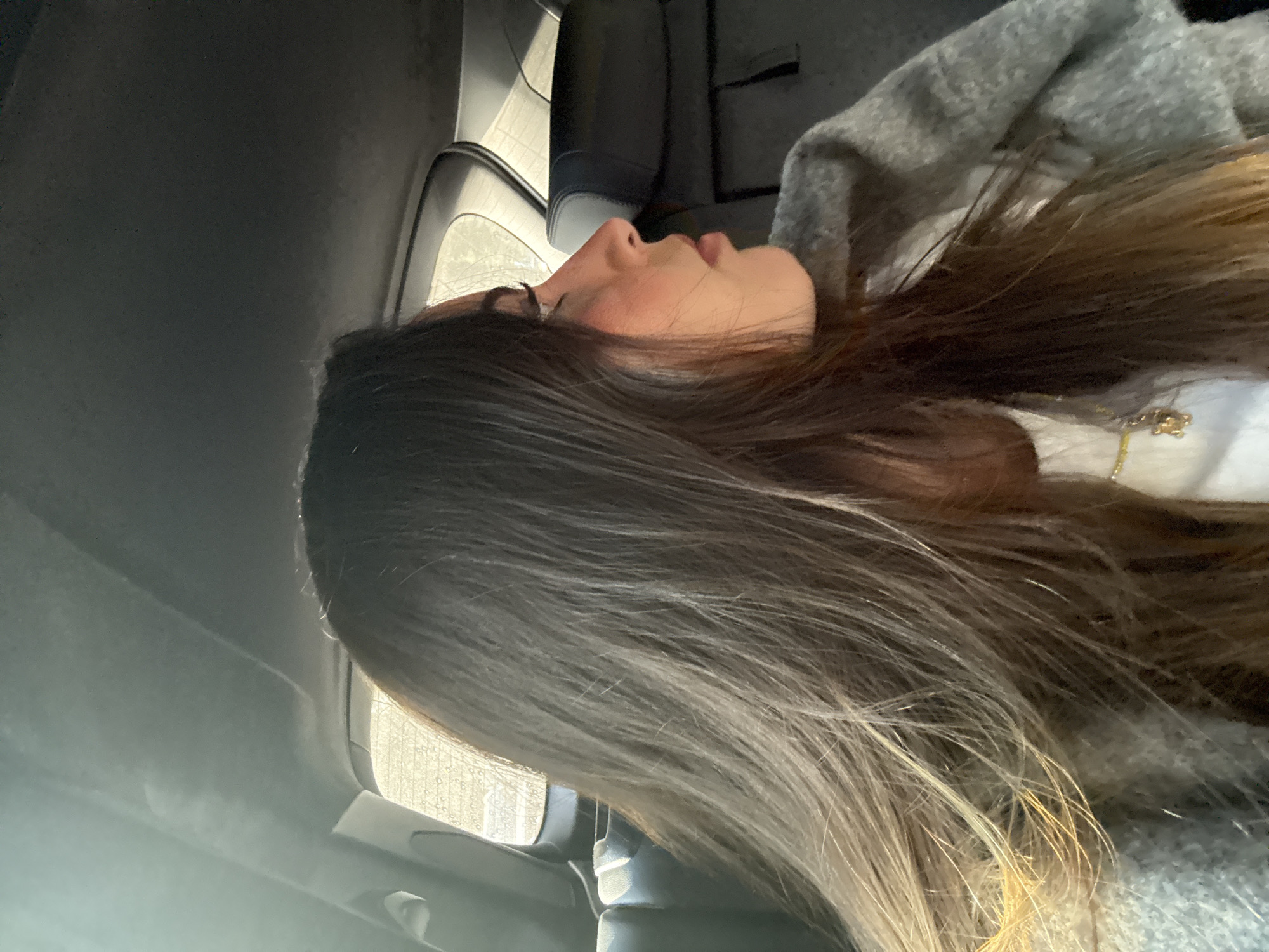 A woman with long, ombre hair lies in the back seat of a vehicle. The lighting suggests it is daytime. The interior of the car is visible, with the car seat and door frame in view. The woman is wearing a light-colored shirt and a gray sweater. The exact location in the city of Puebla, Mexico is not discernible.