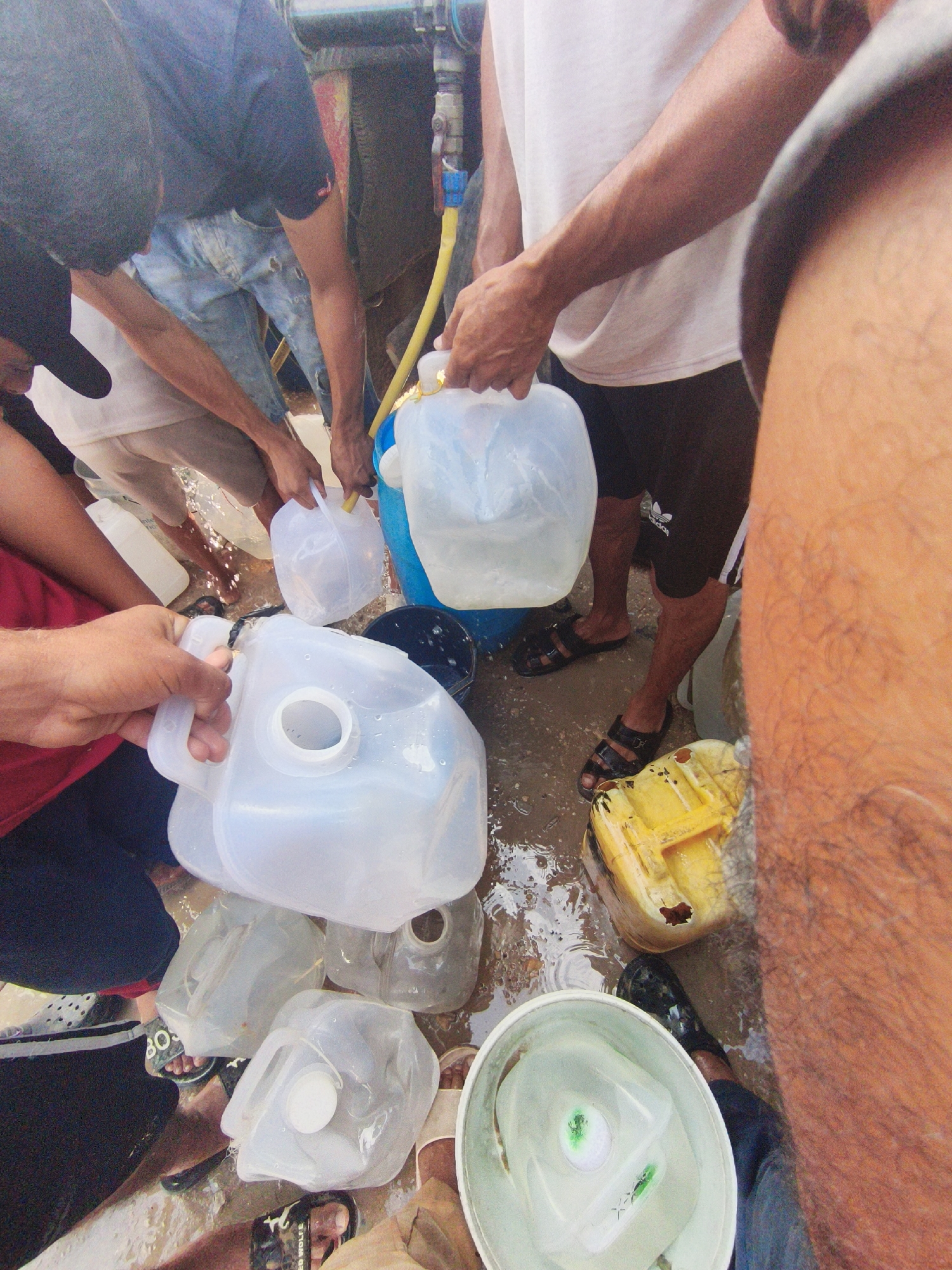 The image shows a group of individuals filling multiple plastic jugs with water from a hose. The scene appears to be outdoors on a wet, muddy surface, possibly a street or a courtyard in an urban environment. Several people are visible, some in focus and others partially obscured, all engaged in the activity of collecting water. They are wearing casual clothing, including t-shirts, shorts, and sandals. The water collection process involves using a yellow hose that is connected to a larger water source, and the water is being poured into various sizes and types of translucent plastic containers. Some jugs are nearly full, while others are being filled. A blue barrel and a yellow jerrycan are also present in the scene. The ground is covered in water and mud, suggesting a recent or ongoing water scarcity issue or a communal water distribution point. The overall impression is one of a practical, collective effort to secure a vital resource.