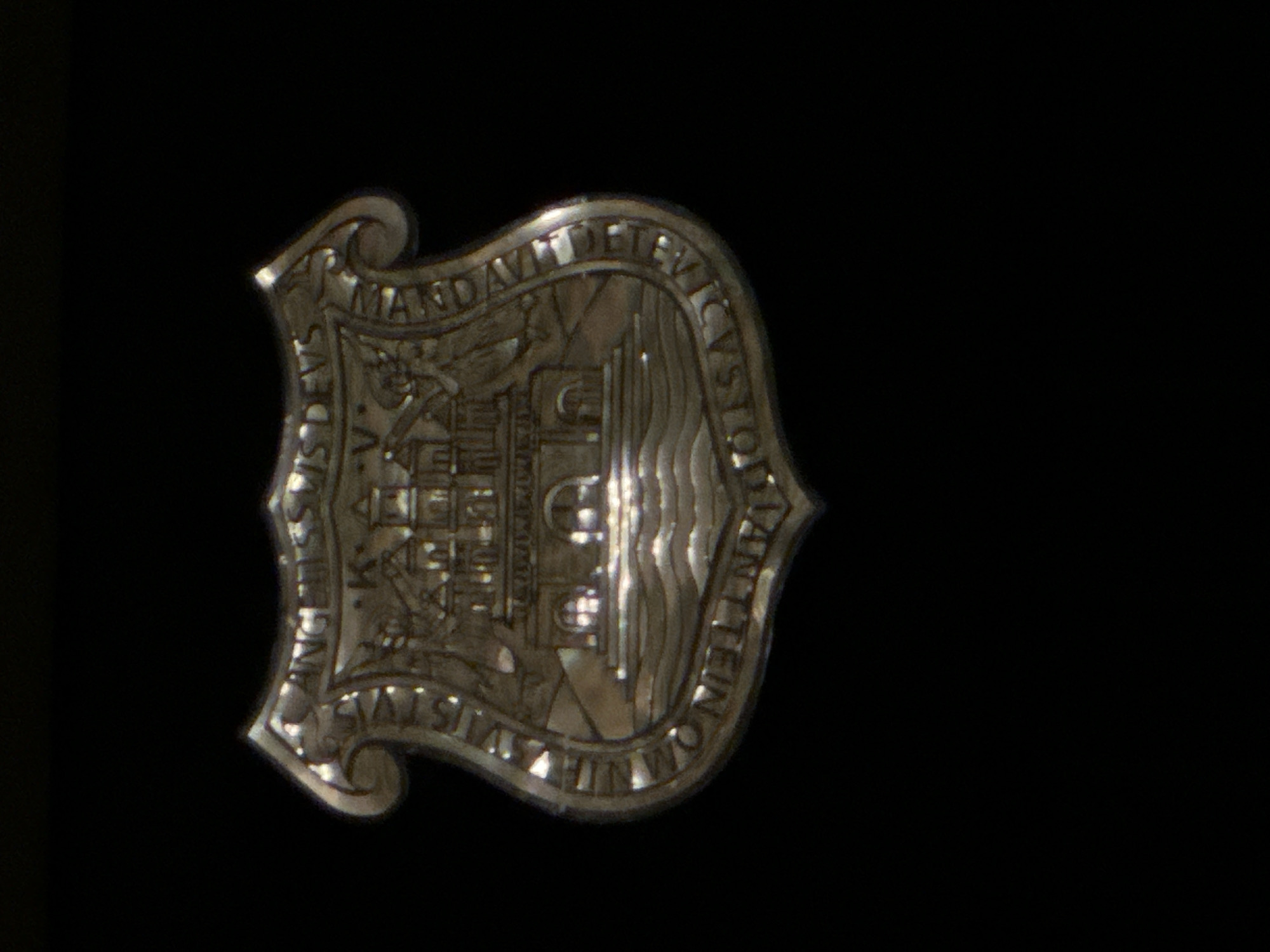 A metallic crest is prominently displayed against a dark background. The crest depicts a building on one side and waves on the other. Text is etched around the perimeter of the crest, with words like "MANDAVIT DEUS CVSTODIS ANG K..." and "VIH OMN STIVS." The location in the city is unknown, as are the location details.