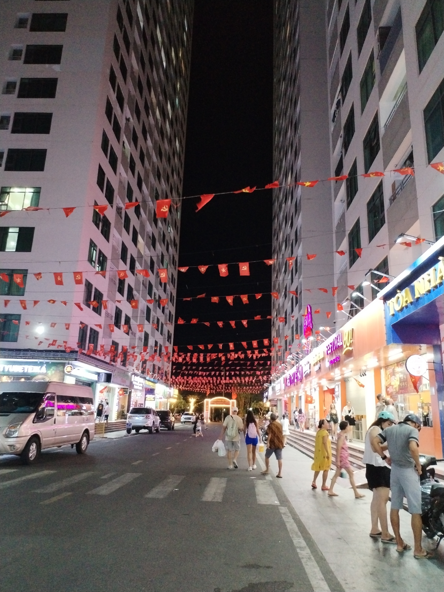 The image shows a street scene in Bac Nha Trang Ward, Vietnam, at night. Two tall, light-colored buildings flank a street decorated with numerous strings of small red flags. Pedestrians walk along the sidewalk on the right side of the street, and some are also on the road. Several vehicles are parked along the road. Stores with illuminated signs line the sidewalk on the right.