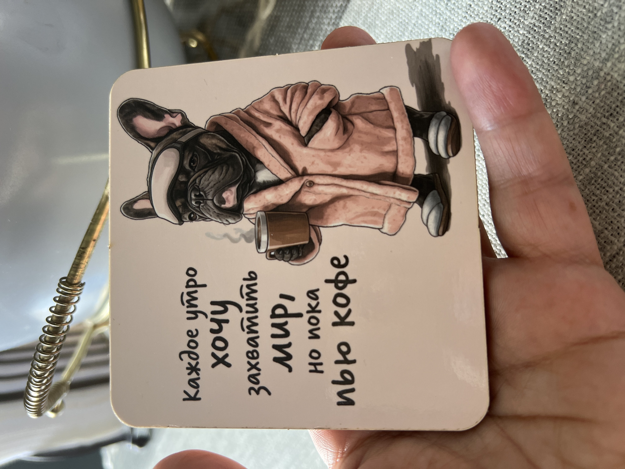 This is a close-up, eye-level, indoor shot of a coaster featuring a drawing of a French bulldog wearing a bathrobe and sleep mask, holding a mug of coffee. The text on the coaster, written in Russian, translates to: "Every morning I want to conquer the world, but for now I drink coffee." The coaster is held by a person's hand, with a textured, light-colored surface visible in the background, possibly a tablecloth. There is no discernible indication of the location being in Mineralnye Vody, Russia; the image solely focuses on the coaster and the hand holding it.