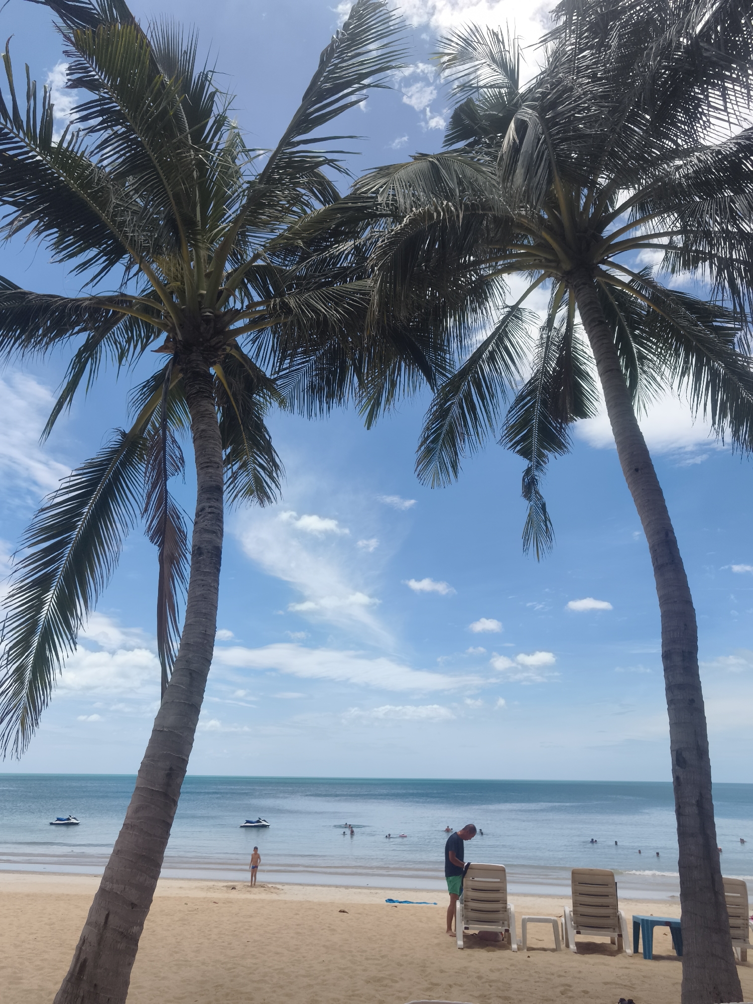 The image depicts a tropical beach scene in the Ko Samui District of Thailand under a bright blue sky with scattered white clouds. Two large palm trees dominate the foreground, their fronds casting shadows on the sandy beach. In the midground, the calm, turquoise ocean stretches to the horizon. Several jet skis are visible on the water, and a few people can be seen swimming or wading. On the beach, a young boy stands near the water's edge, while a man in a dark t-shirt and green shorts stands next to several empty lounge chairs. The overall atmosphere is serene and recreational.