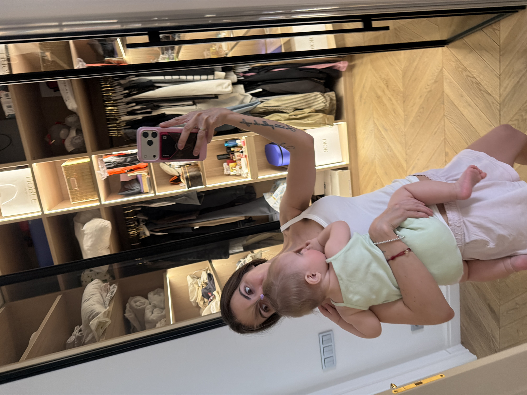 A woman is holding a baby in front of a mirrored closet. The closet is organized and contains folded clothes, handbags, and boxes, some marked "DIOR". The floor is light-colored wood with a herringbone pattern. The woman holds a pink cell phone to take a selfie. The location is in a room in a house, most likely in Krasnoyarsk, Russia, based on the original request.