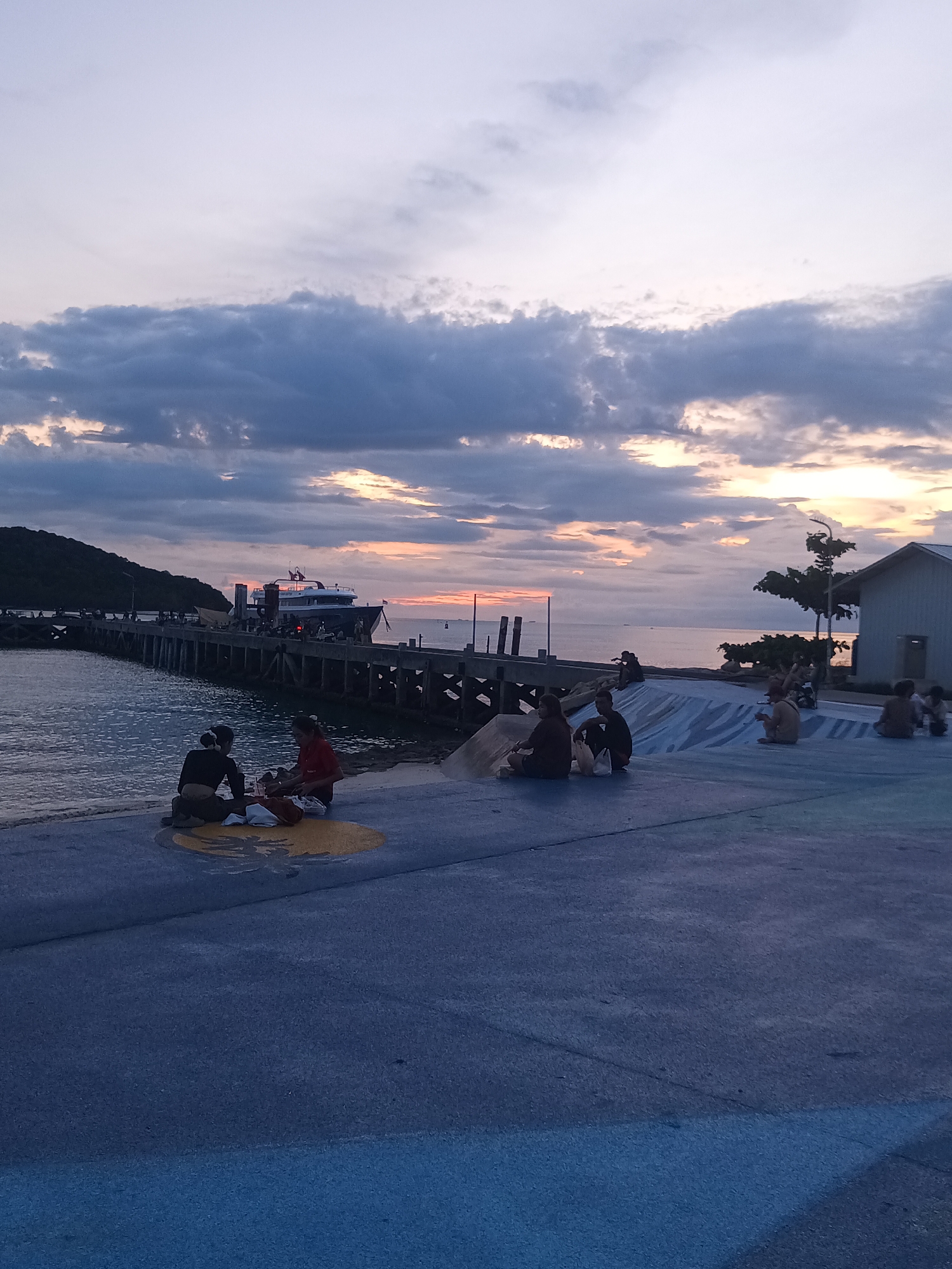 At sunset on the coast of Koh Phangan, Thailand, several people are gathered on a paved area near the water. In the foreground, two individuals sit on a colorful circular marking on the pavement, with bags of belongings beside them. Further back, a group of people are spread out on the paved surface, some sitting and others appearing to be engaged in conversation or relaxation. A pier extends into the sea, with a large ferry docked at its end. The sky is filled with dramatic clouds, with hints of orange and yellow from the setting sun. A small building with a white facade and a simple roof stands on the right side of the image. The overall scene depicts a peaceful evening gathering by the sea.