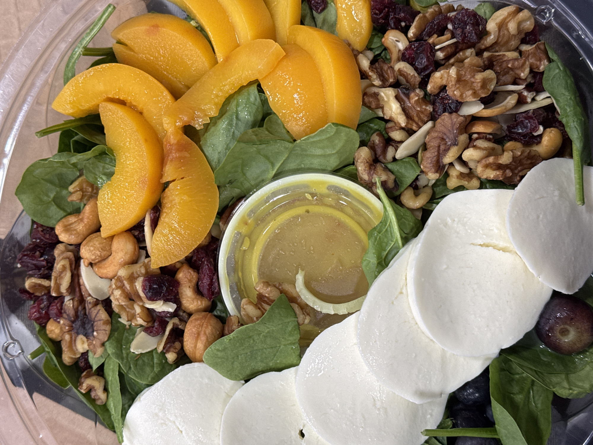 A top-down view captures a salad in a clear plastic container. The salad contains spinach as a base, topped with sliced peaches, a mixture of walnuts, cranberries, and cashews, a small container of salad dressing, and sliced mozzarella cheese. Some blueberries and grapes are visible near the mozzarella. The container is on a light brown surface. There is no evidence to suggest that this image was taken in San Bernardino Tlaxcalancingo, Mexico.