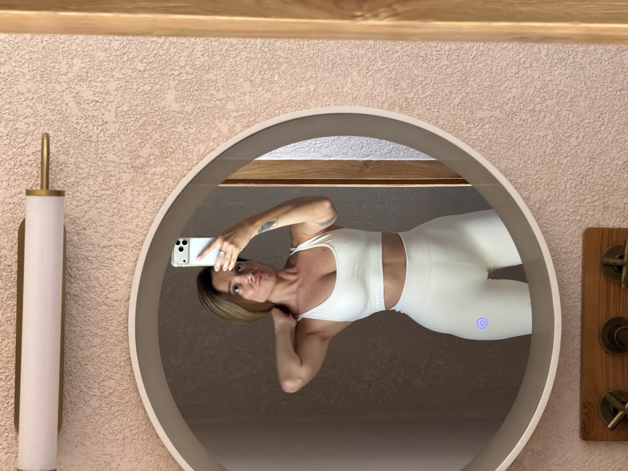 A woman stands in front of a round mirror, taking a selfie with her phone. She is wearing a white sports bra and white leggings, with a glowing blue icon on her left thigh. Her hair is blonde and shoulder-length, and she is making a pouty face. The mirror reflects her body and a portion of the room, which appears to be a bathroom with textured pink walls and a wooden door frame. The image does not contain any identifiable landmarks or elements that would indicate a specific location within the city of Mengwi, Indonesia.