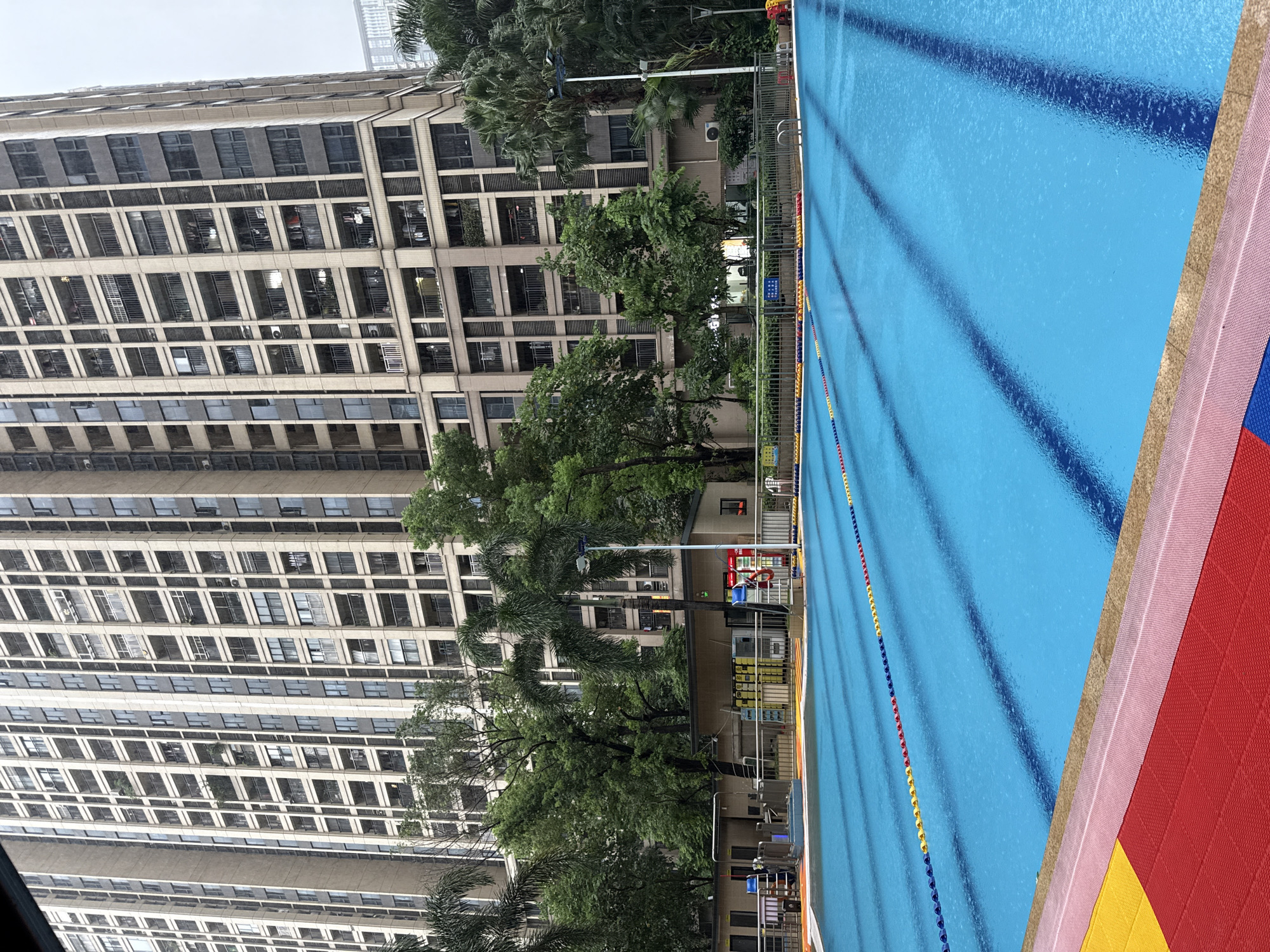The image shows an outdoor swimming pool in Haizhu District, China. The pool is rectangular with several lanes marked by buoys. The water is a vibrant blue, and there are ripples on the surface, suggesting it may be raining or windy. The pool area is surrounded by a fence, and beyond that, there are trees and a tall apartment building. The building appears to be modern, with many balconies and windows. There are also some smaller structures near the pool, which might be changing rooms or equipment storage. The overall scene is one of urban recreation, with the pool offering a contrast to the dense residential architecture in the background.