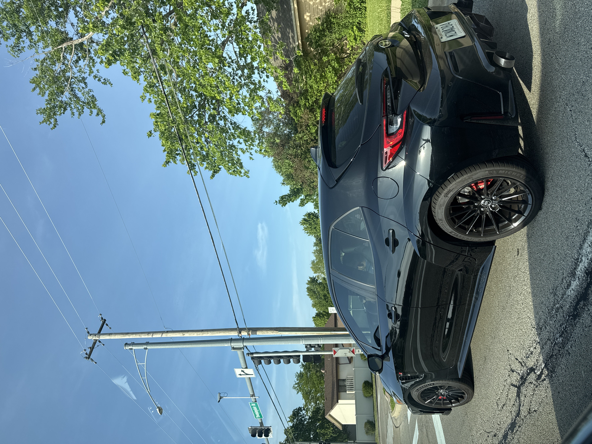 A black hatchback is parked near the corner of the intersection of Bloomingdale Road and W Army Trail Road in Bloomingdale, Illinois. The car has a spoiler and distinctive tail lights. The street sign at the intersection reads "Bloomingdale Rd" and "W Army Trail Rd." A power pole and overhead wires are visible, as are the traffic lights at the intersection. The sky is blue with a few clouds. Trees line the street. The license plate number is N74LB84 and the registered state is Wisconsin.