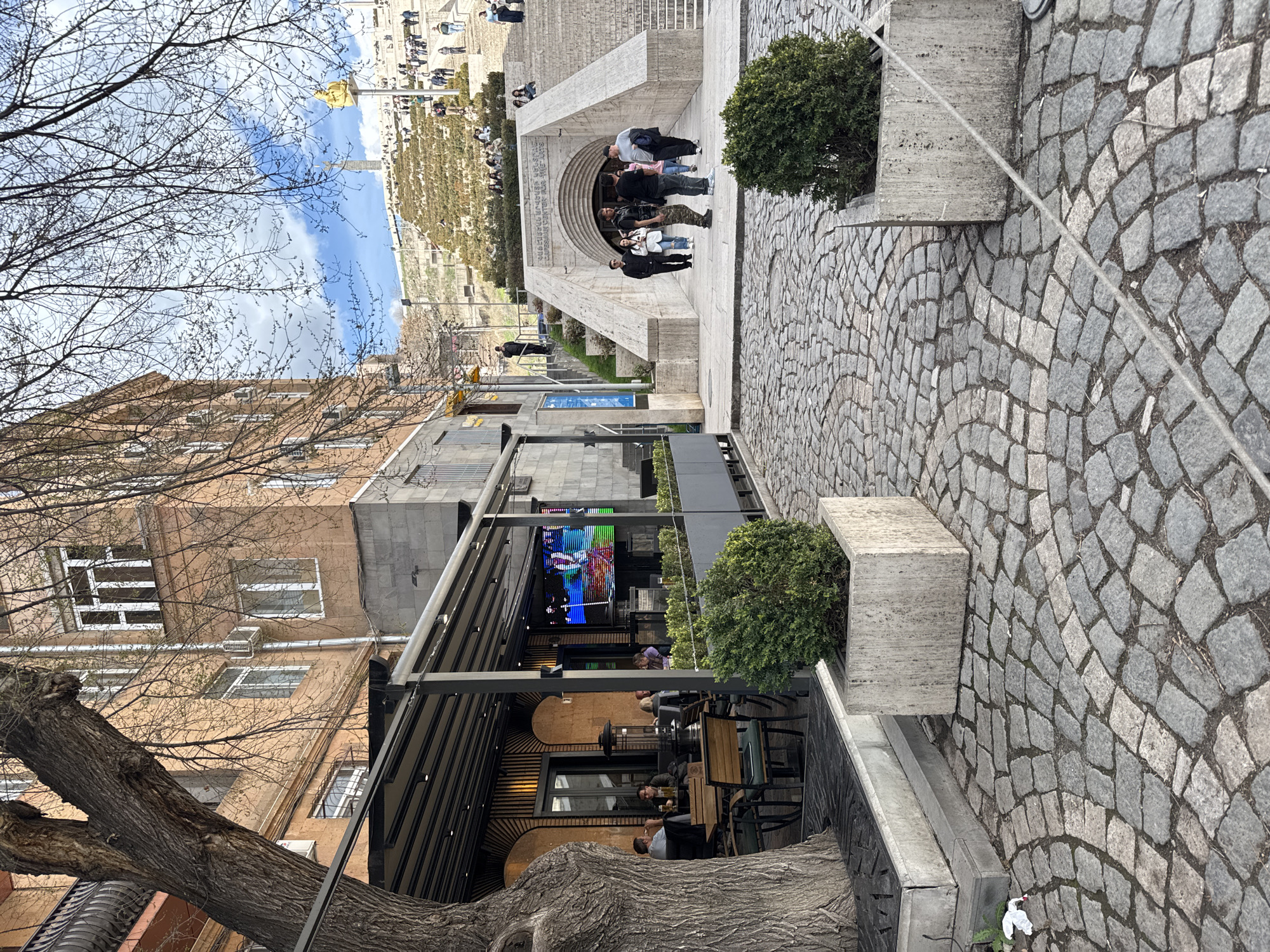 The image depicts a scene in Yerevan, Armenia, showcasing the Cascade complex. The view looks up from a lower level towards the monument's staircase and the surrounding area.
Foreground: A stone-paved walkway occupies the right side of the frame. A planter box containing a round shrub sits along the edge of the walkway.
Midground: A modern outdoor cafe area is seen along the left. It has a retractable awning and a large television screen displaying colorful images. People are seated at tables, seemingly dining or conversing. A large tree trunk is visible on the lower left, partially obscuring the building facade.
Background: The Cascade complex itself is visible in the upper portion of the image. A staircase ascends toward a higher point. Several people are walking on the stairs and gathered at the entrance. A hillside is visible beyond the staircase. The sky is partly cloudy. A street lamp is visible on the left.
