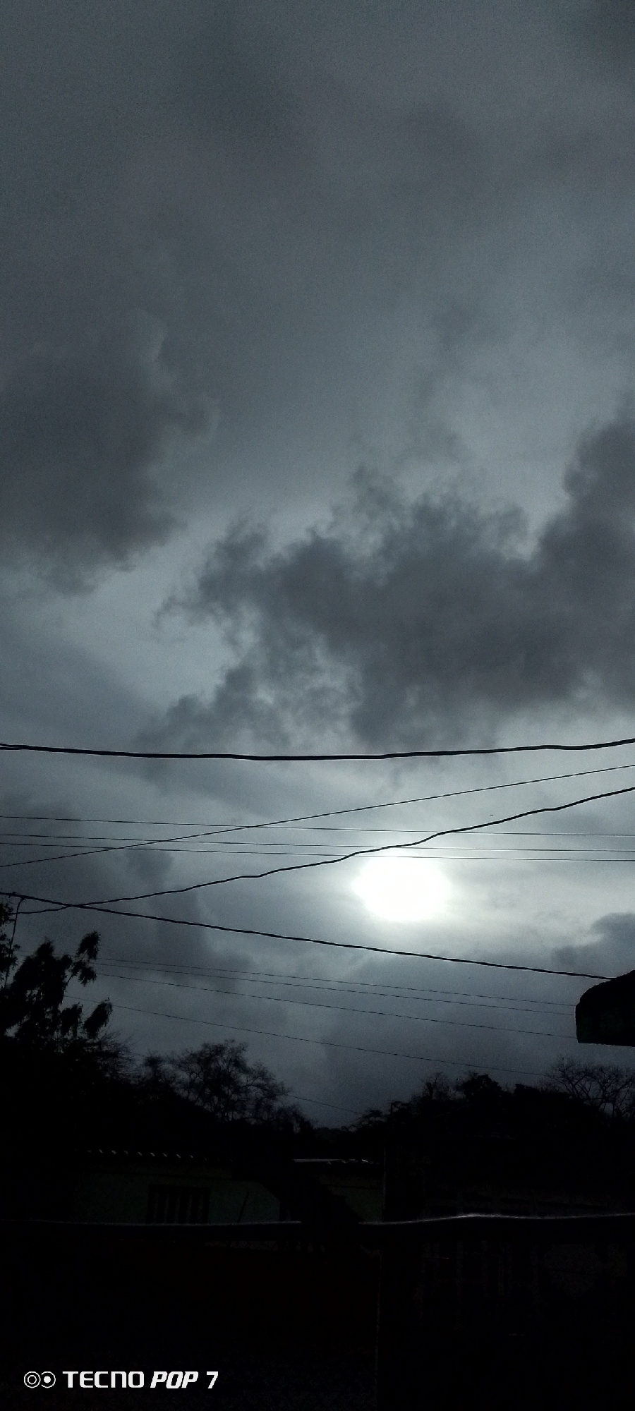 The image depicts a very dark and overcast scene in Altagracia, Venezuela. The sky is filled with heavy, gray clouds. There is a bright area suggesting the sun is trying to peek through the clouds. Several power lines are strung across the mid-ground. The foreground consists of silhouetted trees, foliage, and what appears to be a portion of a building. The scene is very dark, making it difficult to discern specific details of the environment. The TECNO POP 7 watermark is visible in the bottom left corner.