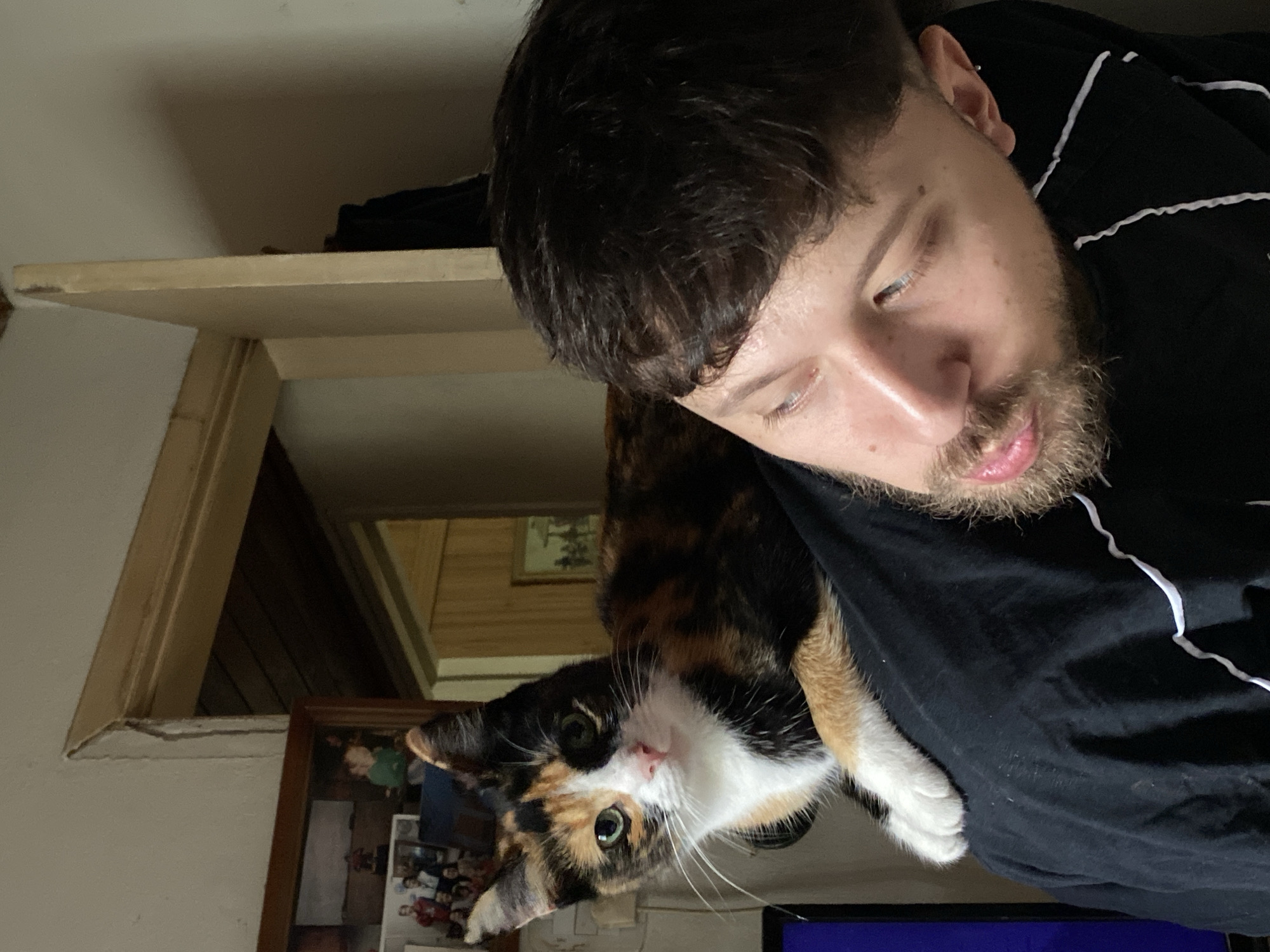 The image presents a close-up, high-angle shot of a man and a calico cat sharing a tender moment in an indoor setting, likely a home in Monte Grande, Argentina. The man, seen from the chest up, has dark hair and a light beard, and is wearing a black shirt accented with white wavy trim. His expression is relaxed, with his gaze directed downwards or to the side, suggesting a moment of quiet contemplation or affection.
Draped over the man's chest and shoulder is a striking calico cat, identifiable by its patches of black, orange/brown, and white fur. The cat's head is turned directly towards the camera, showcasing its bright green eyes and prominent white whiskers, giving it a curious and engaged look. One of its white paws is visible, resting gently on the man's shirt.
The background elements indicate a home environment. A light-colored shelf or wooden surface is visible on the left side, and further back, architectural details like wooden paneling or trim line the walls. A framed picture containing several smaller photographs can also be seen in the lower left background. A hint of a blue screen, possibly a monitor or television, is visible in the bottom right corner. The overall scene conveys a sense of warmth and companionship between the man and his pet. There is no visible text in the image.