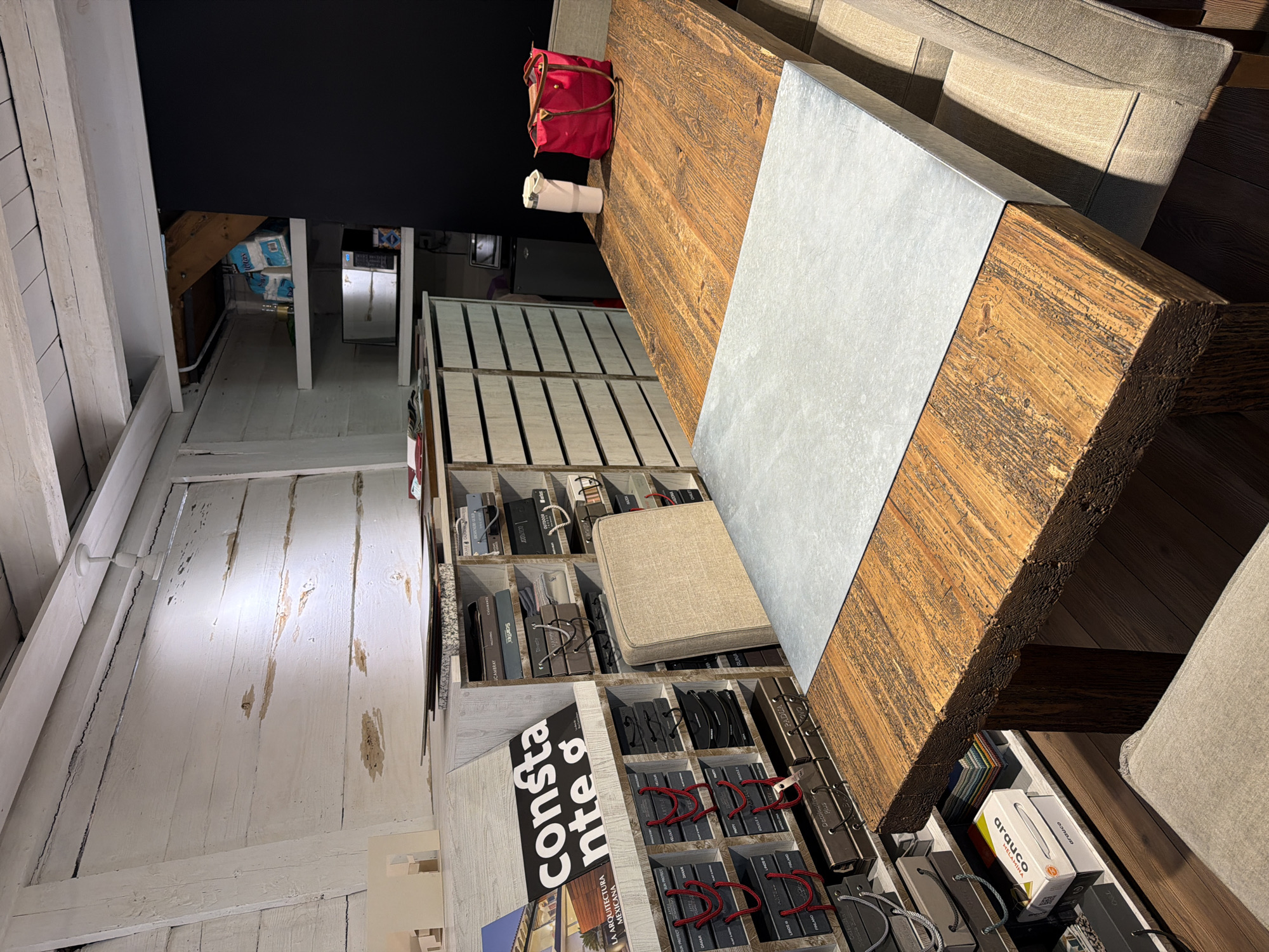 Here is a description of the image:
This indoor shot presents a collection of objects within what appears to be an interior design or furniture showroom, possibly located in Puebla, Mexico due to Spanish text on product packaging. The scene is brightly lit, with a contrast between the white-painted wooden plank walls and ceiling and the dark background in one corner.
Dominating the center is a large, wooden table with a light gray, possibly zinc, inlay running down the center. The table appears to be of rustic design with a thick, roughly finished top. Partially visible are sections of a beige sofa near the table.
Below the table and to the left, a shelved display unit contains various product samples, including leather swatches, fabrics, and product packaging. Brands such as "arauco," "ScanTex," and "Decoleather Plus" are visible. A black and white display card with the word "constante" is prominent on one of the lower shelves.
A red backpack and a rolled-up item (possibly a towel or fabric) are positioned on the table. The floor is made of dark wood planks. The image is taken from an elevated angle, giving a comprehensive view of the curated display.
