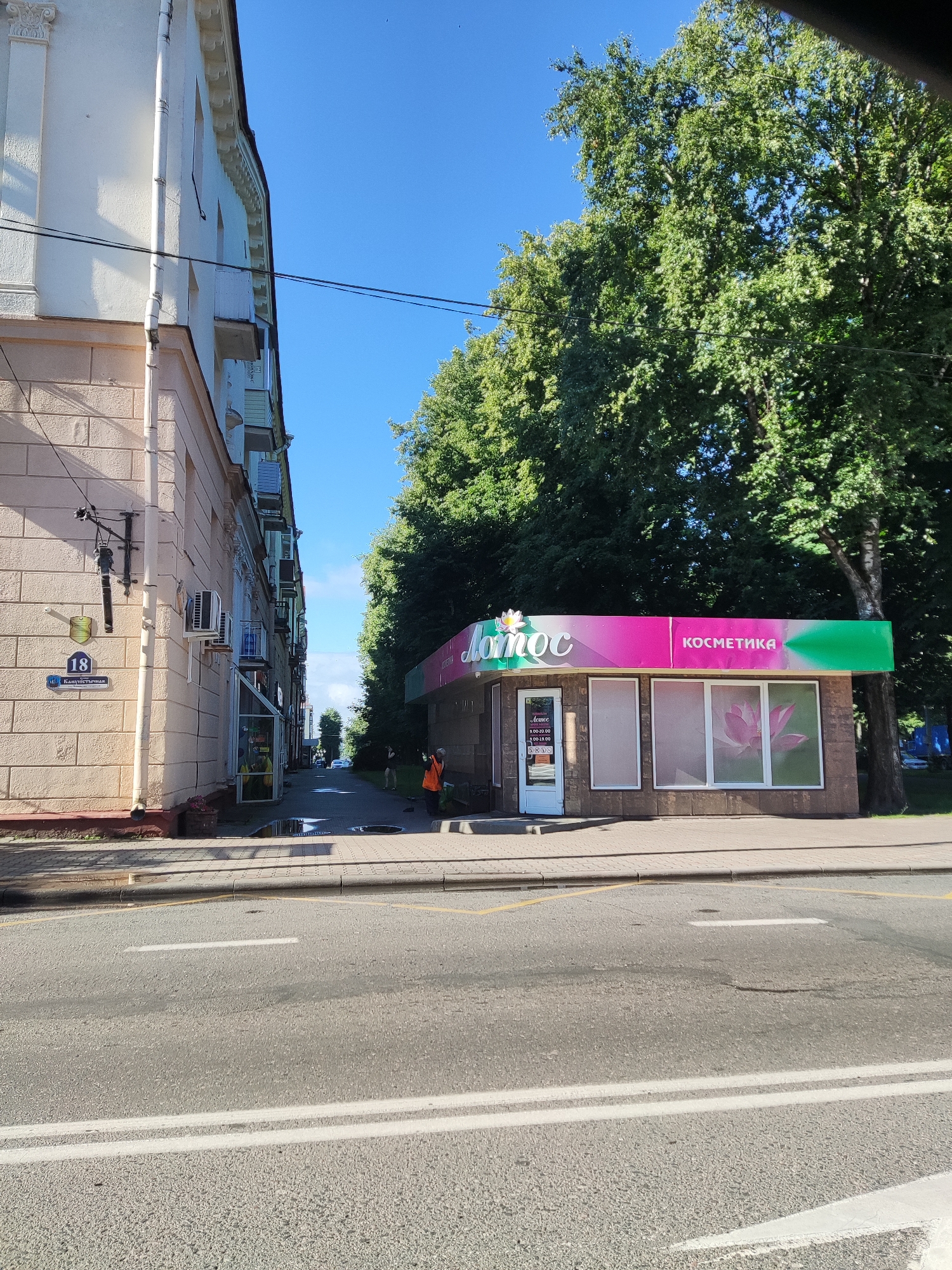This is an eye-level, outdoor shot of a commercial building situated next to a road and a sidewalk. A person in an orange vest is visible on the sidewalk, possibly engaged in cleaning or maintenance. The building features a prominent sign with the word "Лотус" and "КОСМЕТИКА" written in Cyrillic, indicating it is a cosmetics shop. The shop has large windows, one of which displays a pink lotus flower graphic. The scene appears to be taking place during the day, under a clear blue sky. Lush green trees line the right side of the frame, partially obscuring a second building in the background. On the left, a multi-story building with a light-colored facade is visible, showing architectural details and several air conditioning units. A street sign with the number "18" is also visible on this building. The road in the foreground has double white lines and appears wet in patches.