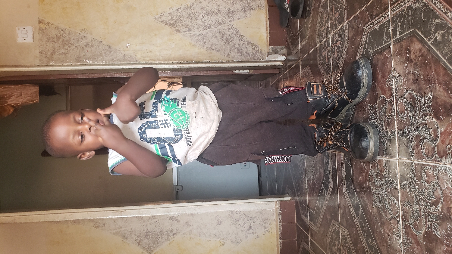 A young child, likely a boy, with dark skin and short dark hair, is captured standing indoors in what appears to be a hallway or entryway in Narayi, Nigeria. He is dressed in a light-colored, short-sleeved t-shirt adorned with green, blue, and black graphics and text that include "PROFESSIONAL," "TRAIN," "Hiya!," and "FREAK!" He wears dark brown or grey long pants, on which the partial text "UNNING" is visible. A notable detail is his footwear: oversized black work-style boots with distinctive orange and brown patterned laces.
The child is facing the camera, making a playful or silly expression by touching the corners of his mouth with his index fingers. The background features light-colored walls, possibly cream or off-white, displaying some textured or distressed patterns. A white electrical light switch is mounted on the wall to the upper left. The floor is covered with ornate brown and black patterned tiles. In the distance, a darker interior space is visible, along with a pair of dark slippers or sandals. The lighting suggests it is daytime. The image captures a candid, lighthearted moment.