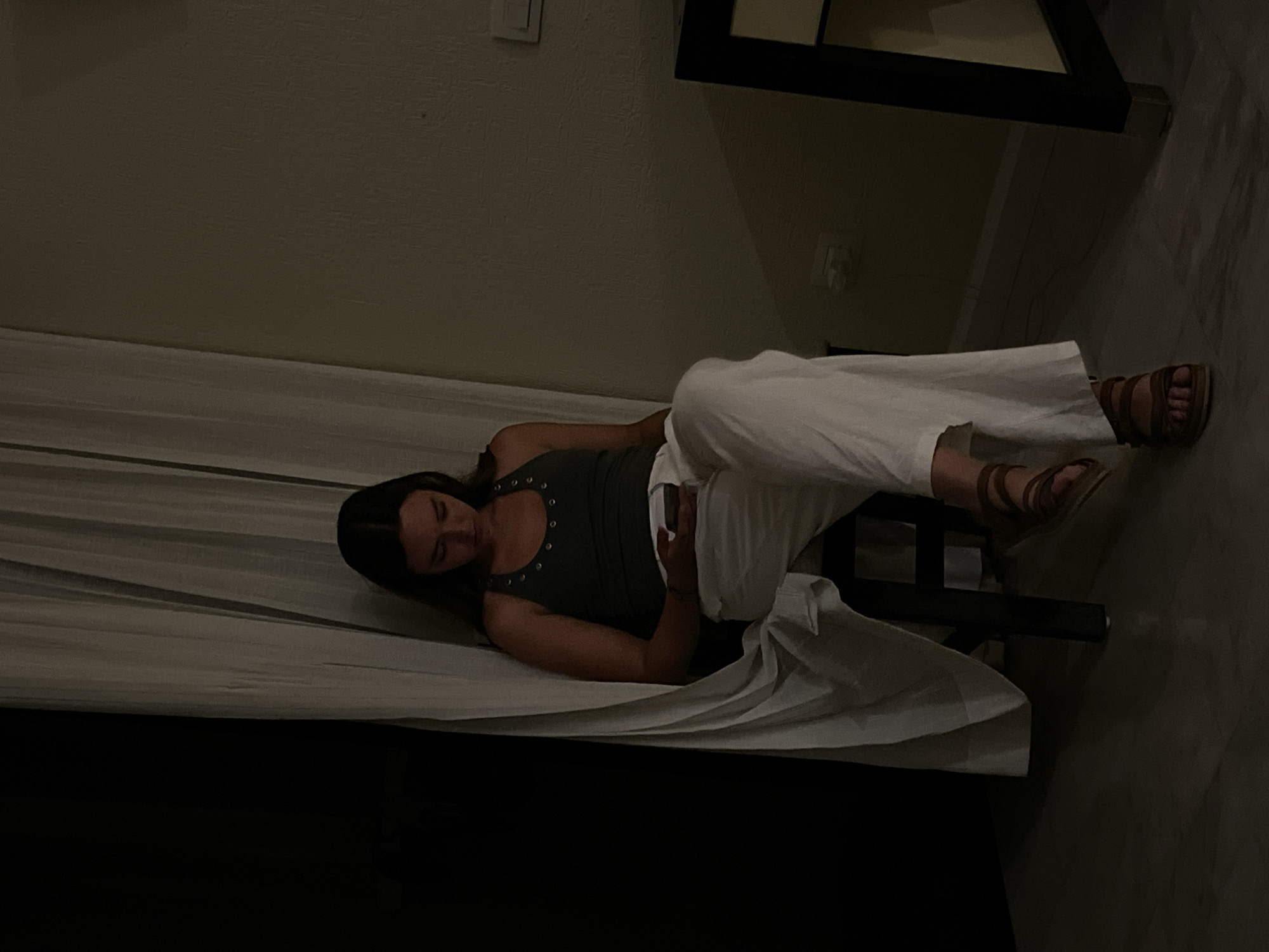A woman is lying on a bed in a room. She is wearing a dark tank top and white pants. She is holding a phone in her hand. Her sandals are visible on her feet. The bed is covered with a white sheet. A black frame is visible in the background. The location is Playa del Carmen, Mexico.