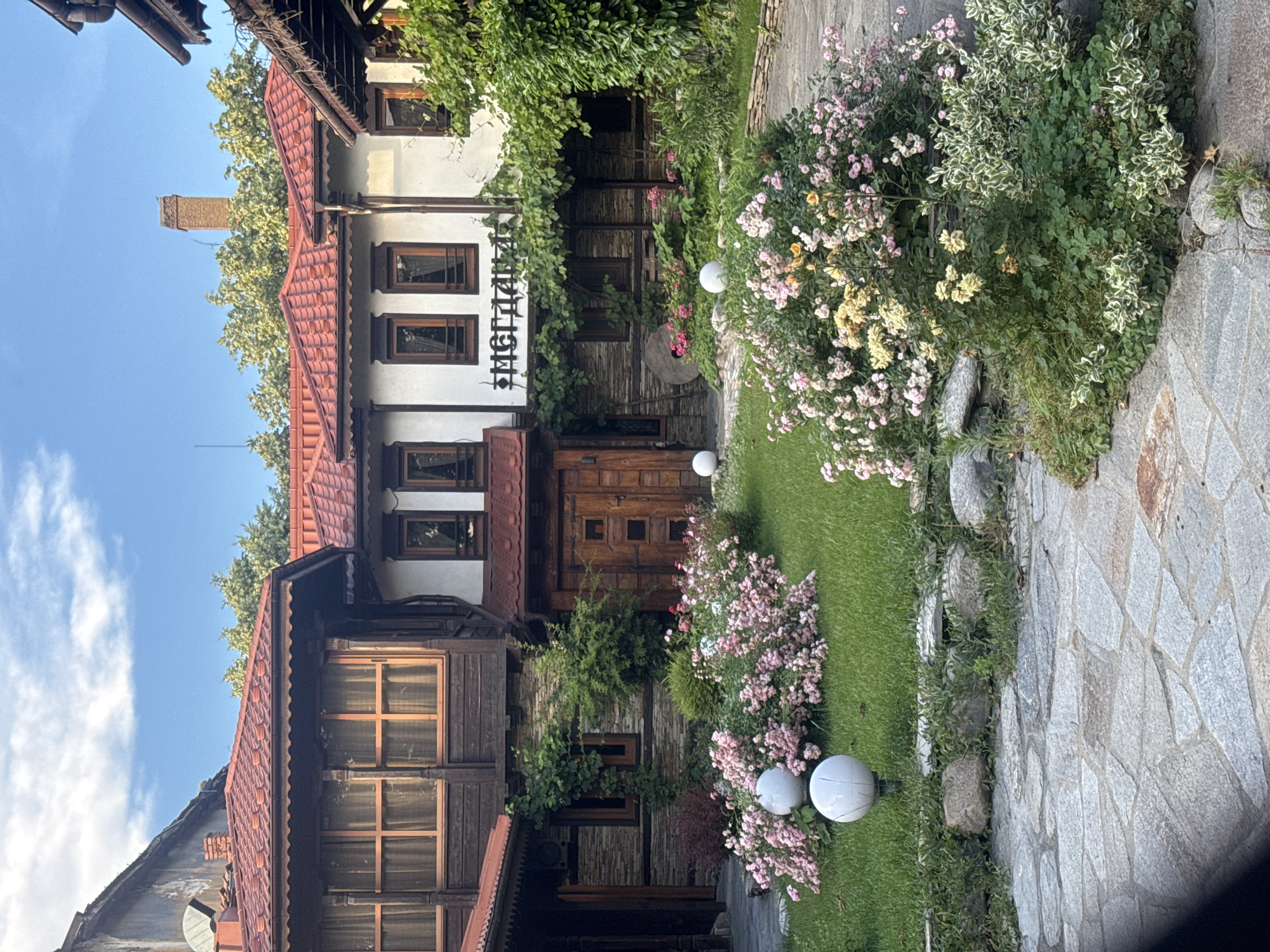 This is a vertical, full shot of a traditional Bulgarian house with an adjacent garden and stone pathway. The house features red tiled roofs, white plastered upper walls, and wooden lower sections with visible timber beams. Multiple windows with wooden frames are scattered across the facades. A chimney is visible above the roofline against a bright blue sky with sparse clouds.
The garden is lush and green, with a vibrant display of pink and yellow flowers. Two spherical white lights are placed in the grass near the flowers. A stone pathway runs along the right side of the image, composed of irregular grey flagstones. The overall setting suggests a warm, sunny day, likely late afternoon or early evening given the light.
Visible text in Cyrillic script reads "ЭМГДАДЖИ" above a wooden door, possibly indicating the name of the establishment or a sign. The scene is picturesque and evokes a sense of rustic charm and tranquility, characteristic of a residential or hospitality setting in Plovdiv, Bulgaria. There are no people visible in the image.