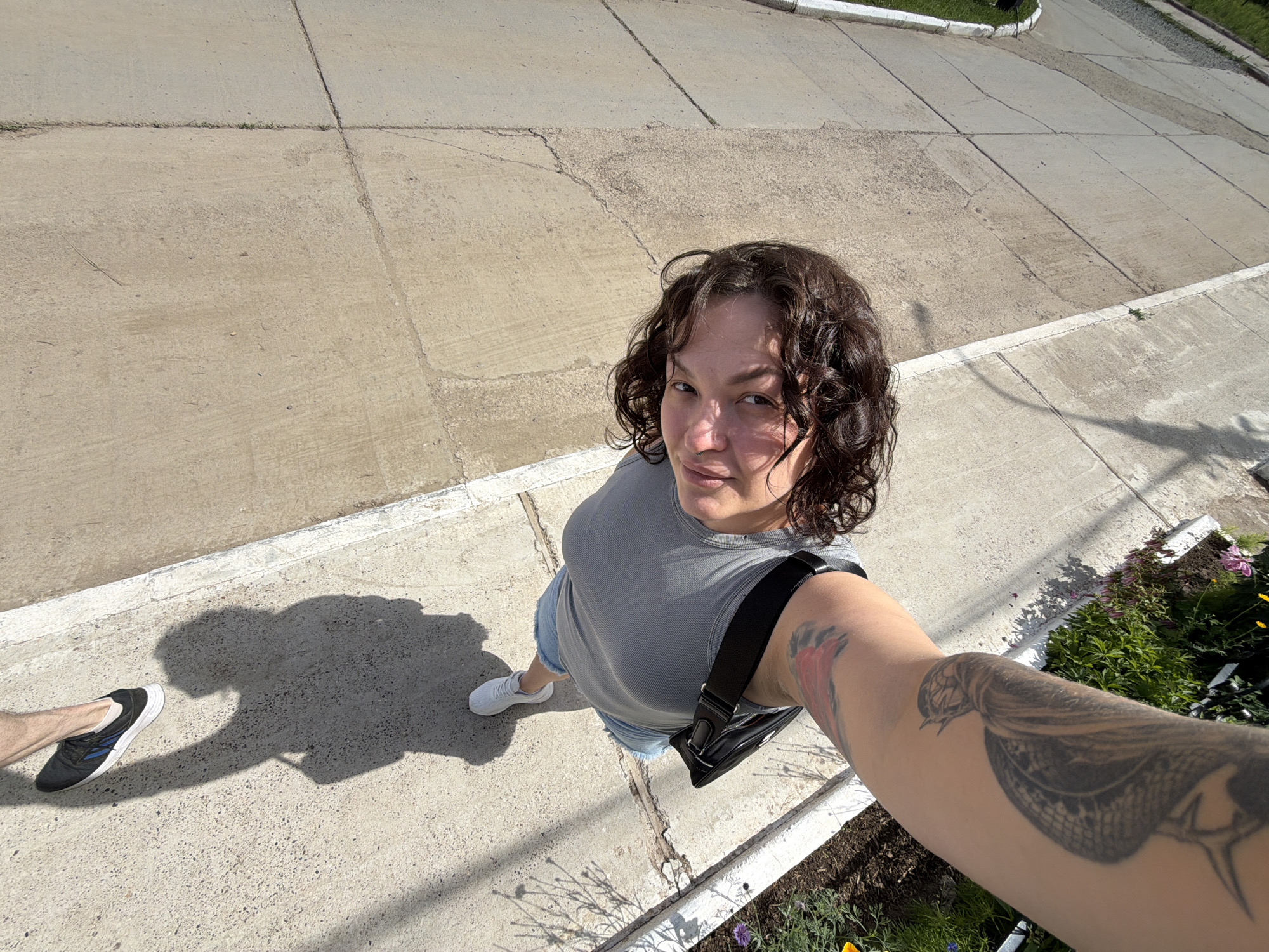 The image shows a woman holding a camera and taking a selfie. She is wearing a gray t-shirt, denim shorts, and white sneakers. She has curly dark hair and tattoos on her arm. The background is a concrete sidewalk with some greenery and flowers. The sun is casting shadows on the ground. The image was taken in Chaikovsky, Russia.