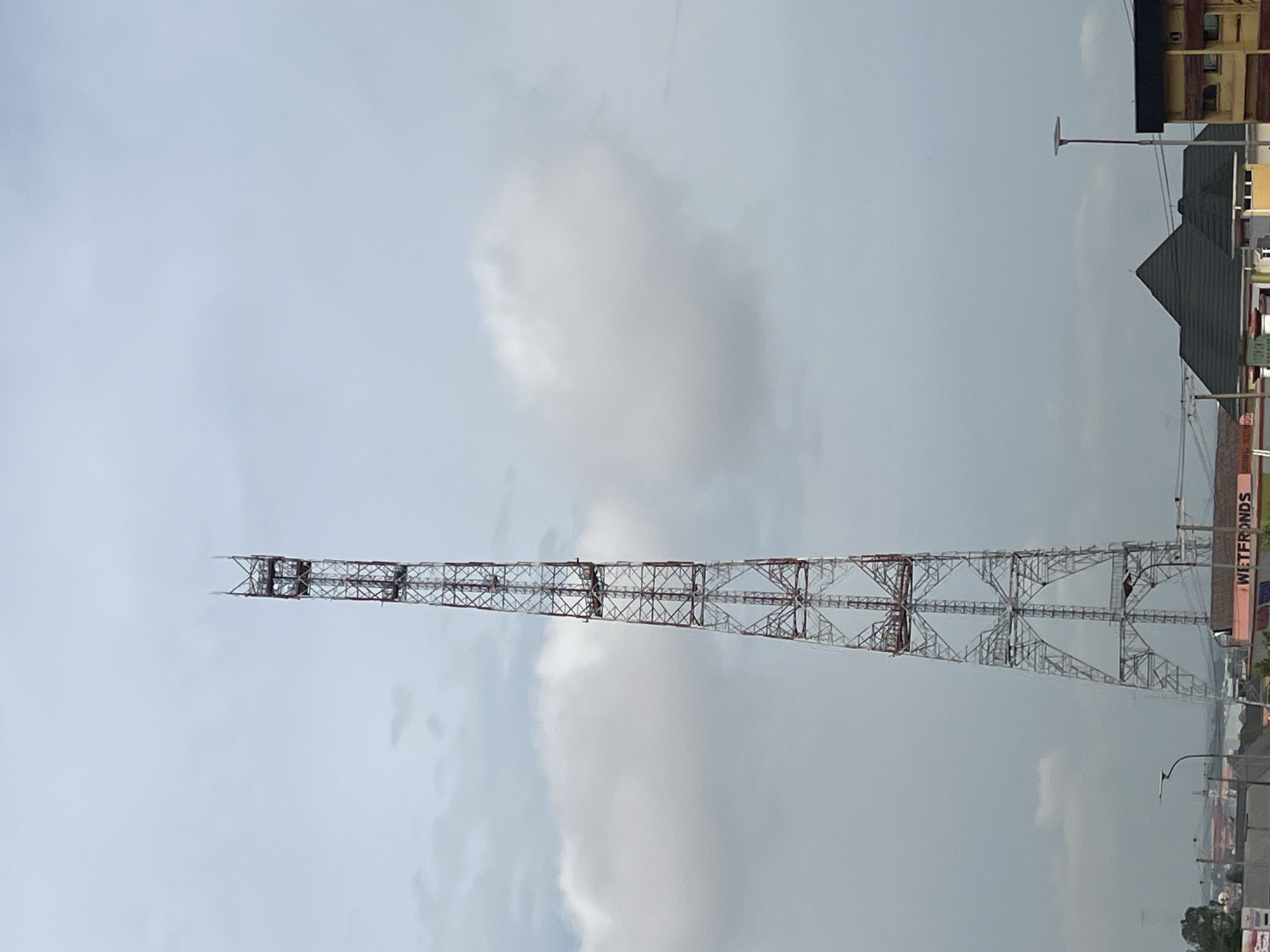 A tall, lattice telecommunications tower with red and white structural elements stands prominently against a cloudy sky in Onitsha, Nigeria. The tower is composed of interconnected metal beams, forming a rigid, triangular framework that tapers towards the top, where it terminates in a pointed structure. Below the tower, a cityscape is visible, with various buildings and streetlights. One building in the foreground features a sign with the text "WETFRONDS" in pink lettering. Trees can be seen on the left side of the image, and a body of water is faintly visible in the distance. The overall atmosphere suggests an overcast day in an urban environment.